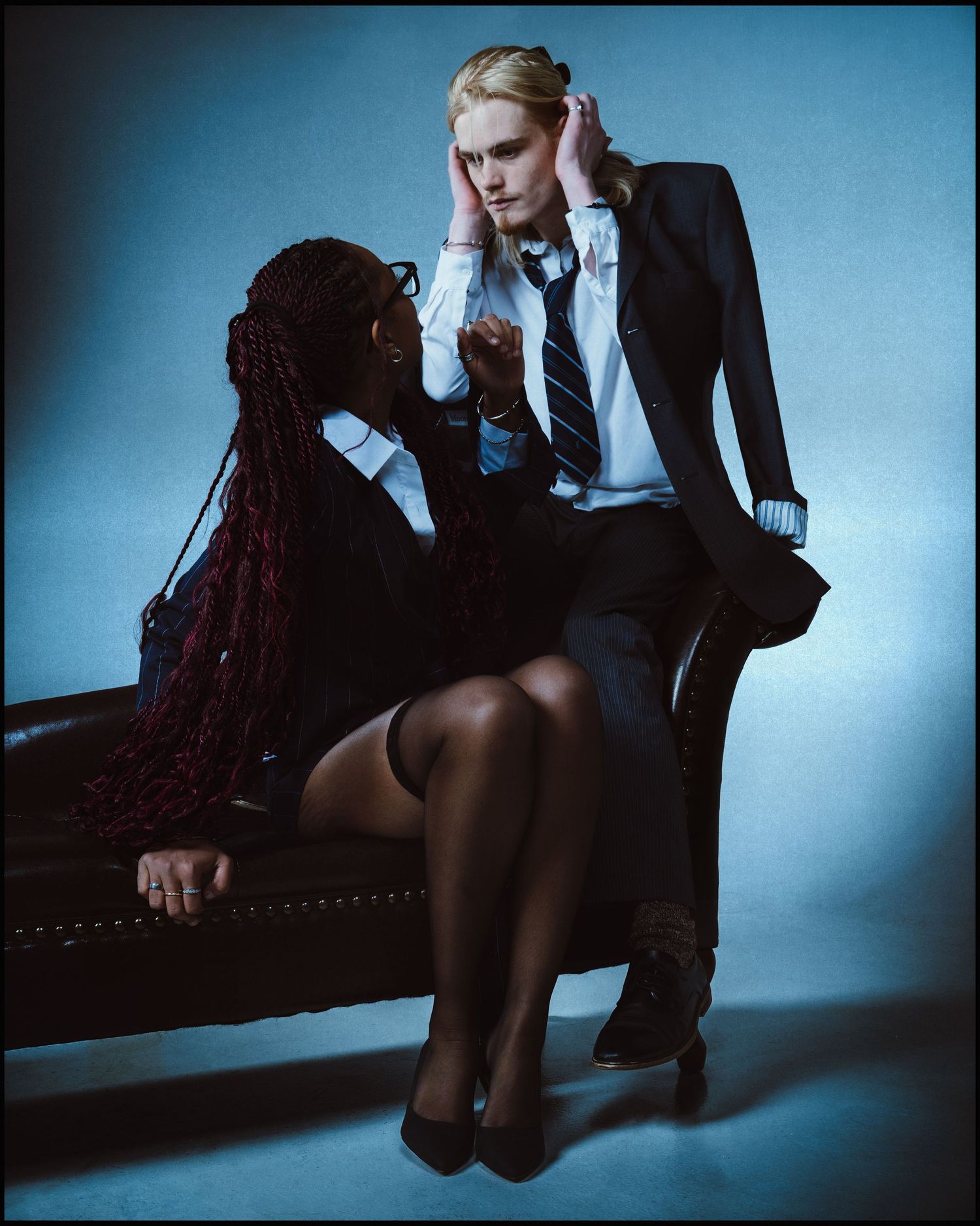 Two women in school uniforms sit together on a leather bench in a moody blue studio portrait.