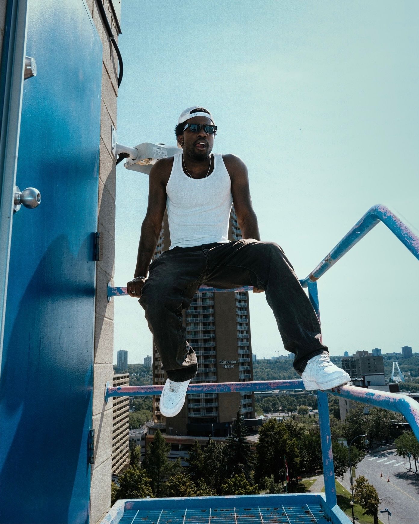 Man in white tank top sitting on rooftop railing with city skyline background.