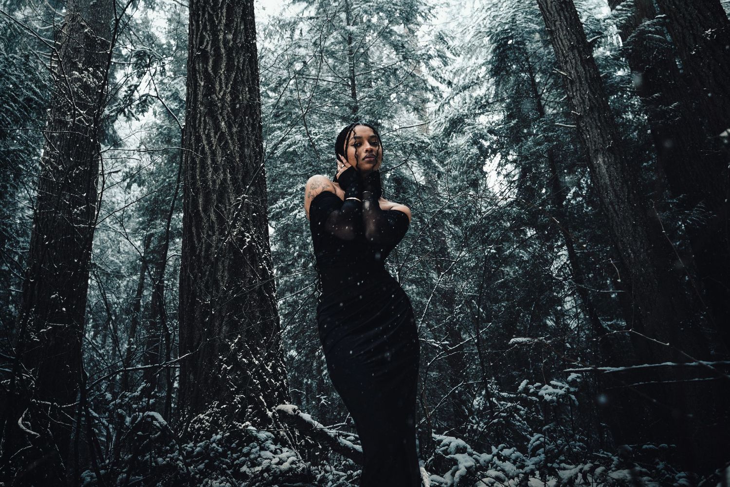 Woman in black dress posing dramatically in snowy forest among tall trees.