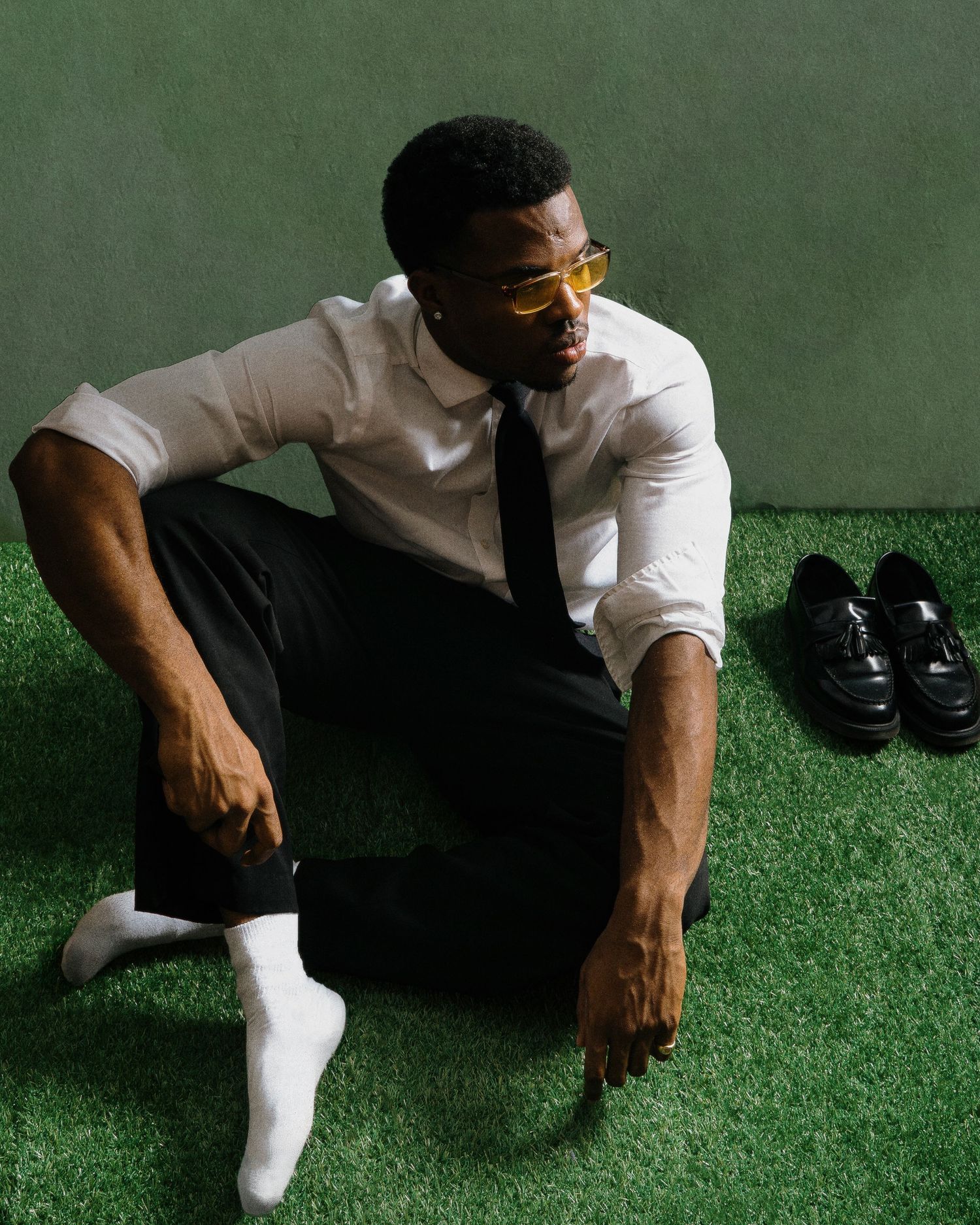 Man in white shirt and black tie sitting on grass with loafers beside him.