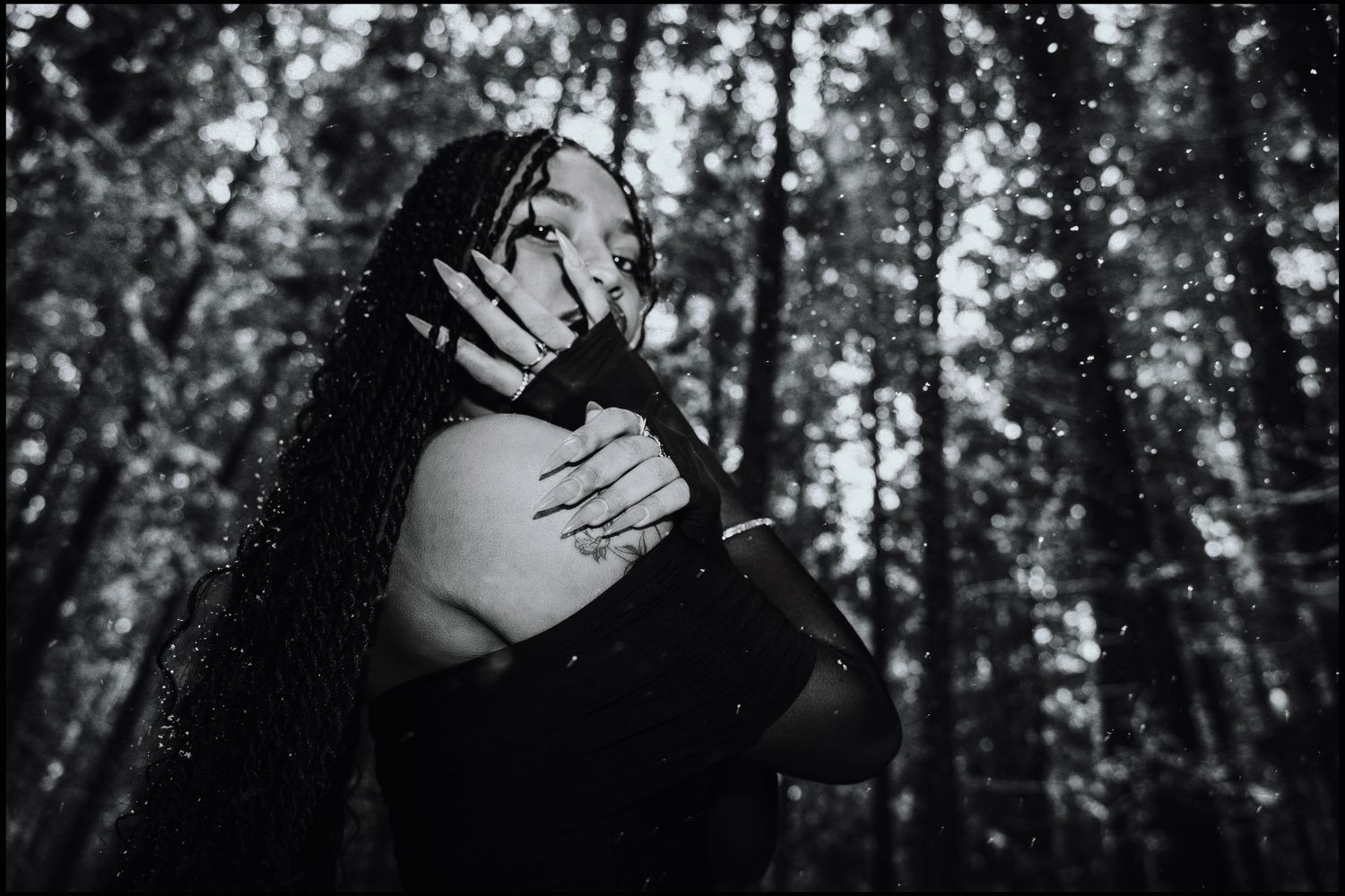 Black and white photo of woman with long braids embracing herself in snowy forest.