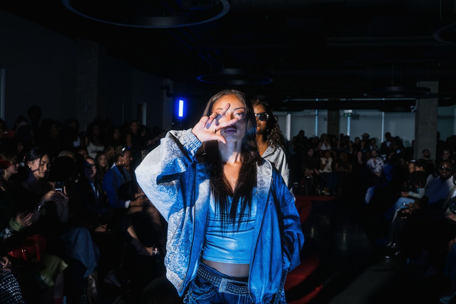 Female model in a blue cropped jacket making a peace sign gesture on a dimly lit fashion runway.