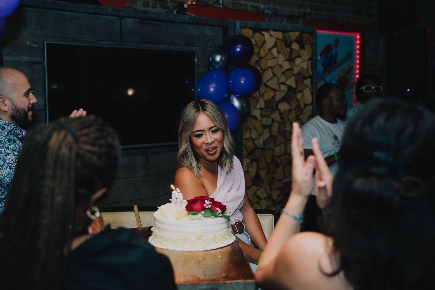 A woman smiles over a white cake decorated with red roses at a birthday celebration with blue balloons.