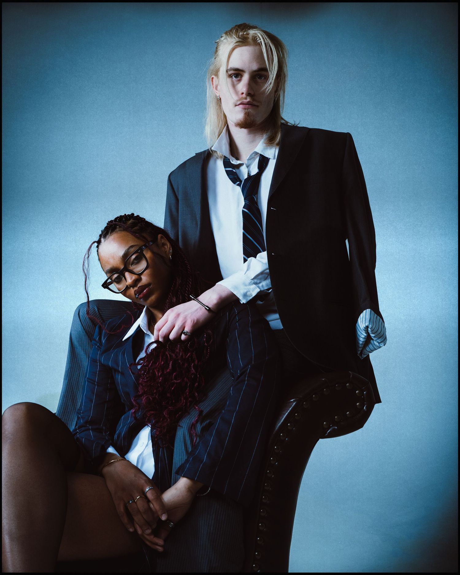 A Black woman and tall blonde man in school uniforms pose together on a chair in a blue studio.