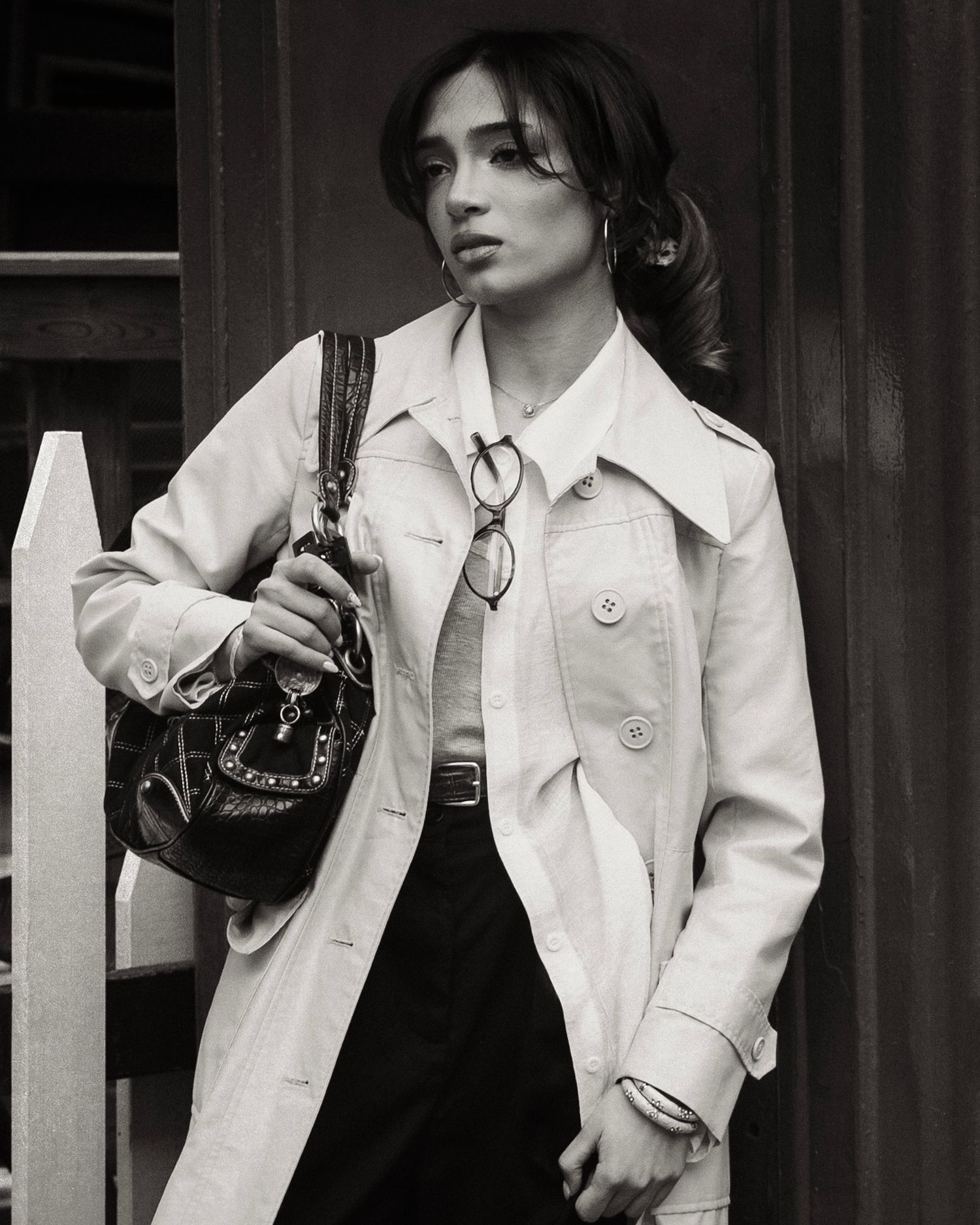 Stylish woman in white trench coat carrying designer bag poses in black and white street photo.