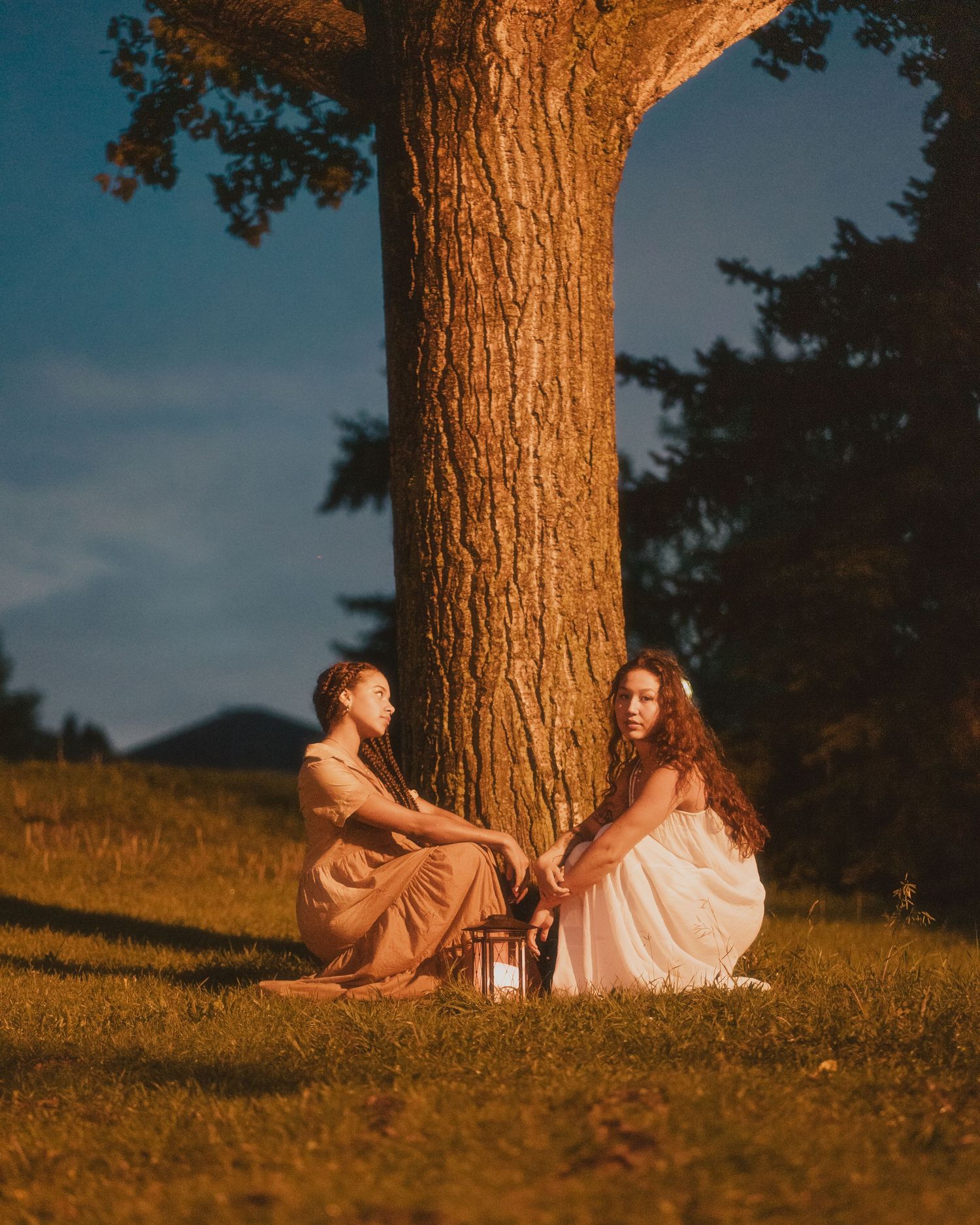 Two women in flowing dresses sit together under a large tree at dusk with a lantern beside them.