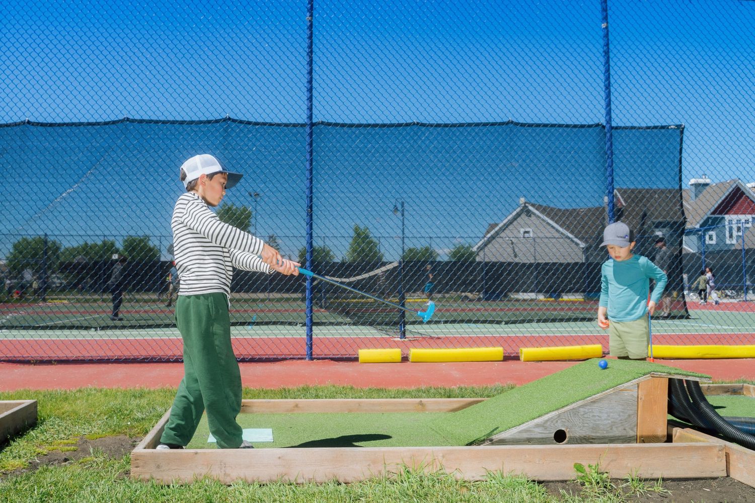 Two children playing mini golf on a sunny day near a tennis court outdoors.