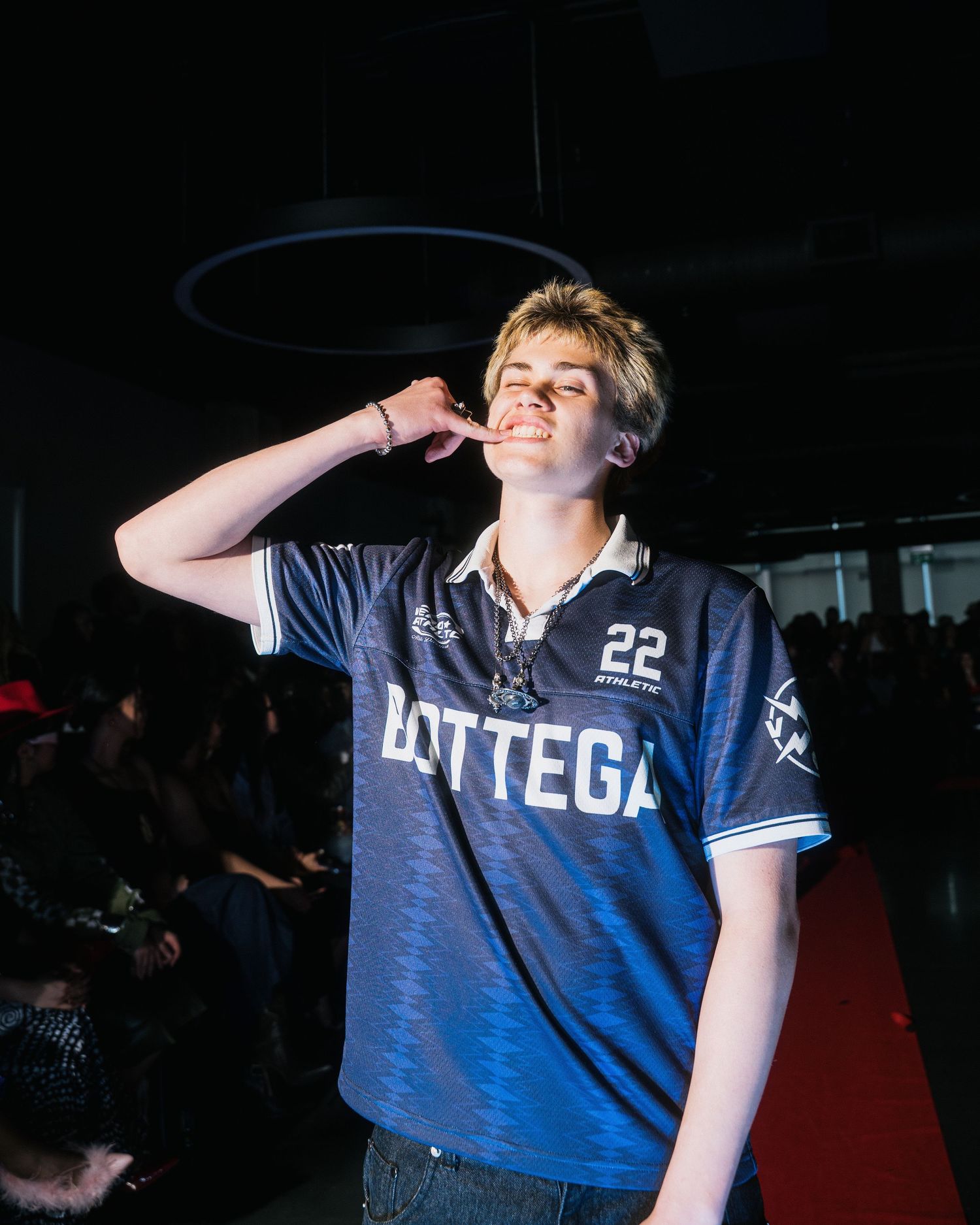 Young man in Bottega number 22 jersey smiles and poses at an indoor fashion showcase.