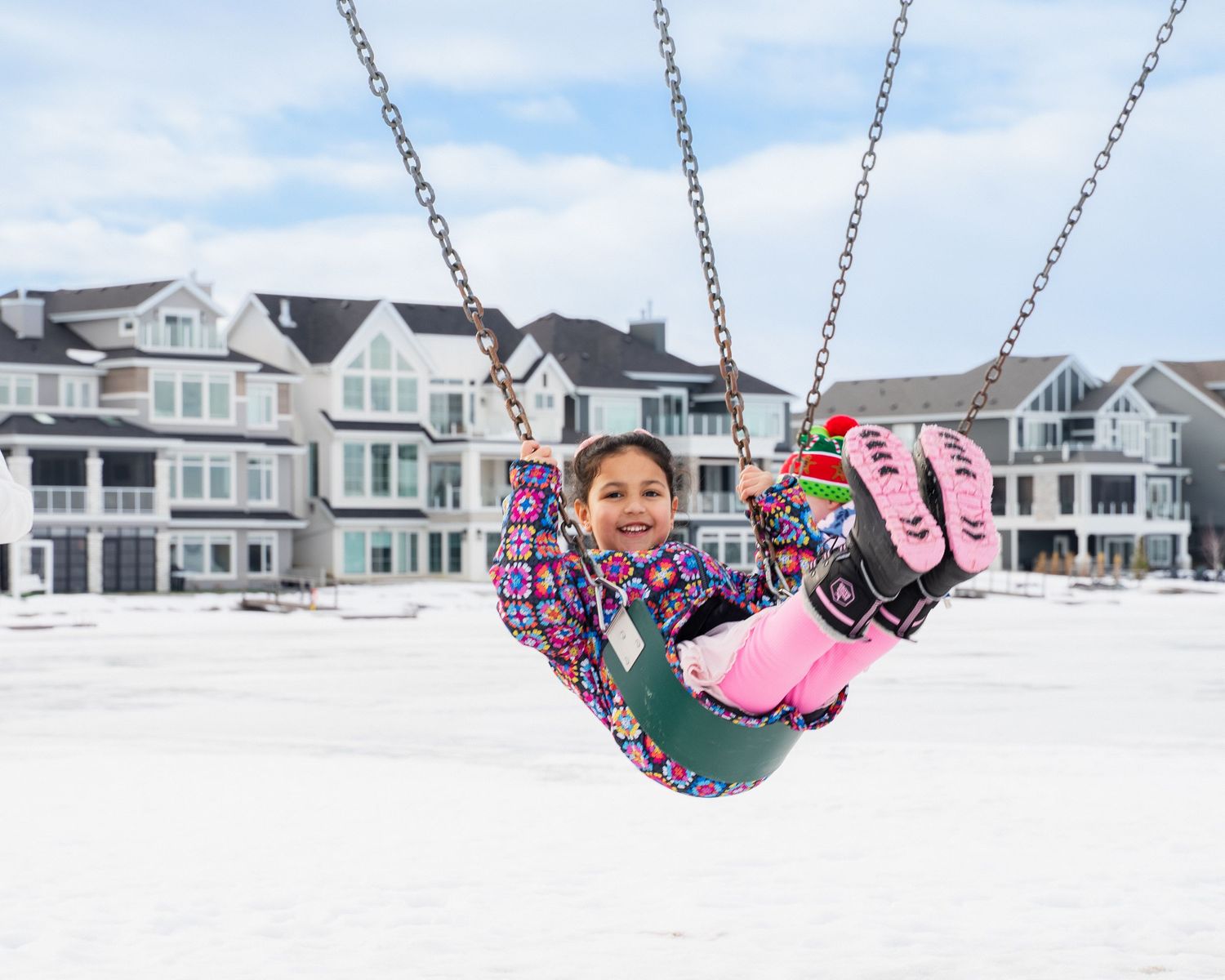 Happy girl in colorful winter jacket swinging on playground swing with snowy townhouses in background.