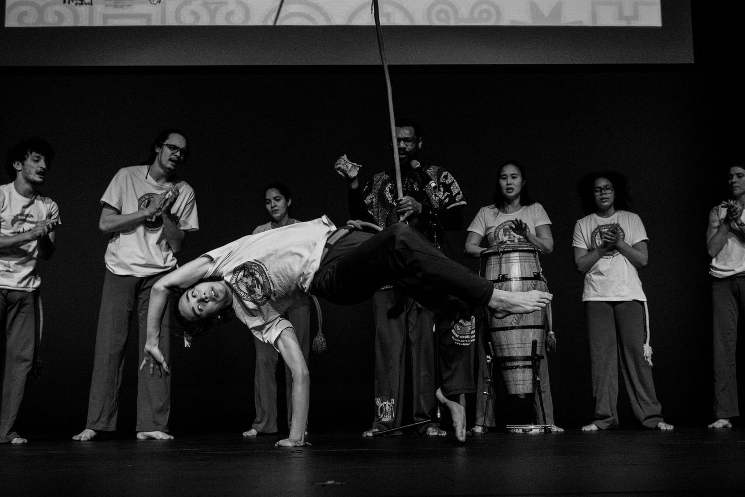 Capoeira performer does acrobatic move on stage with musicians and group members.