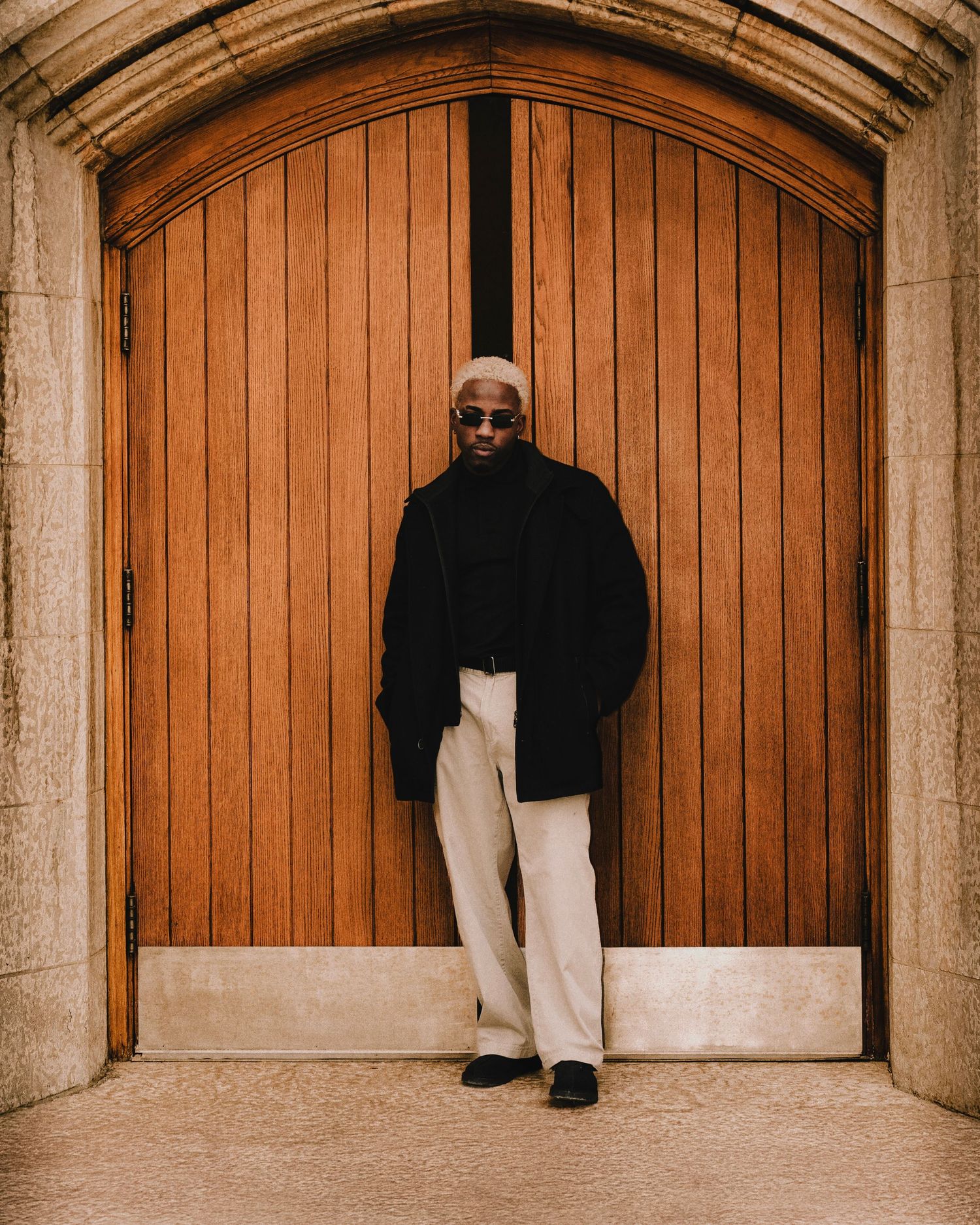 Stylish man in black coat and cream trousers stands before large wooden arched doors.