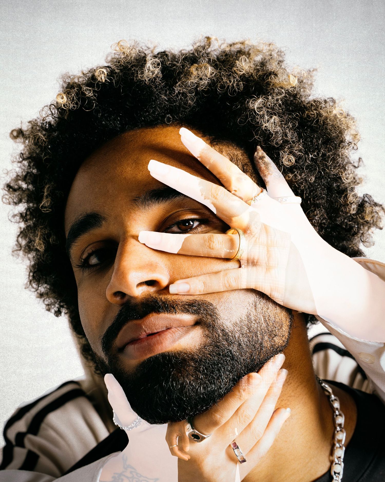 Man with afro and beard holding hand over face wearing chain necklace.