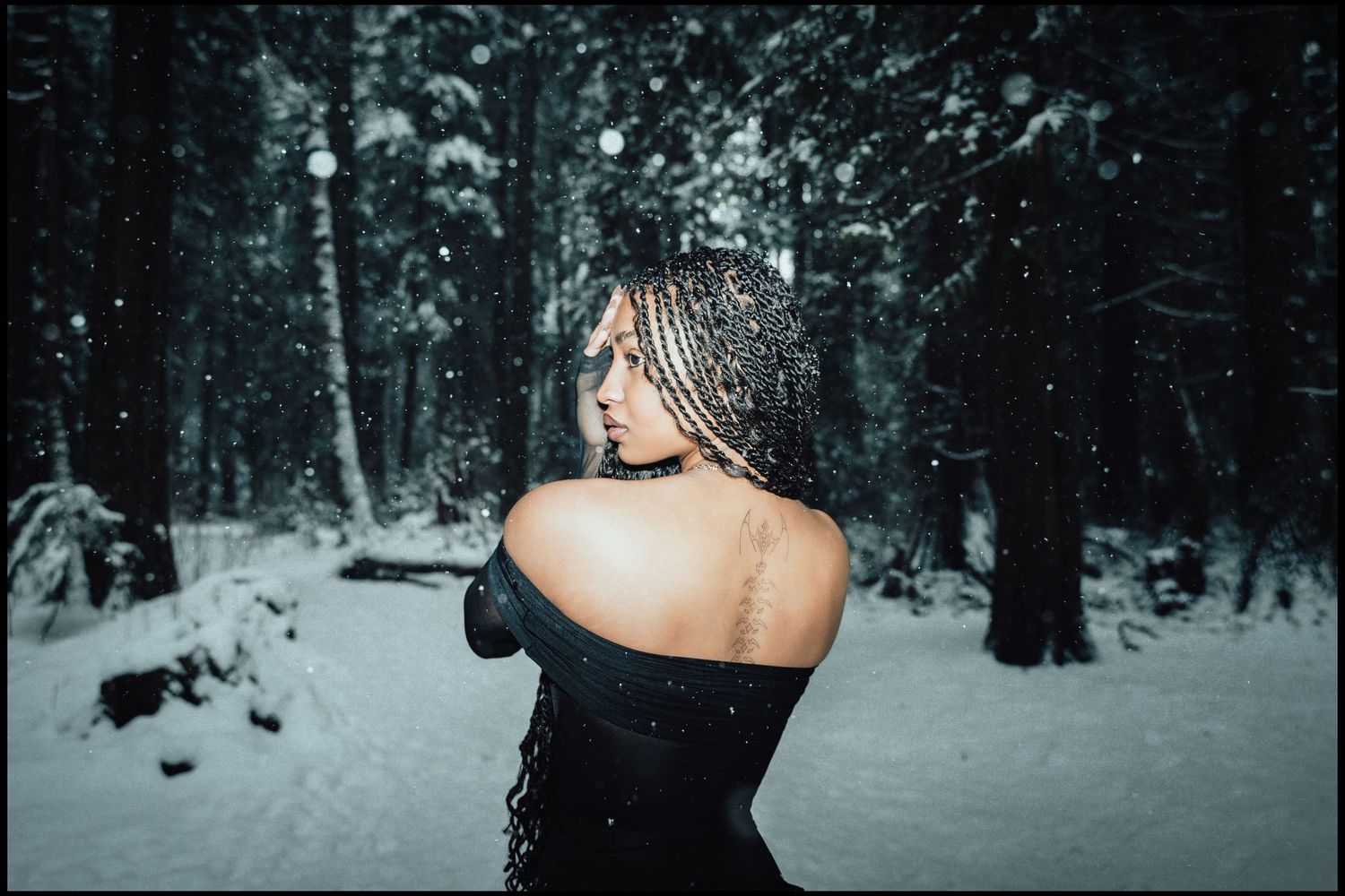 Woman with braids shows shoulder tattoo in black off-shoulder top in snowy forest.