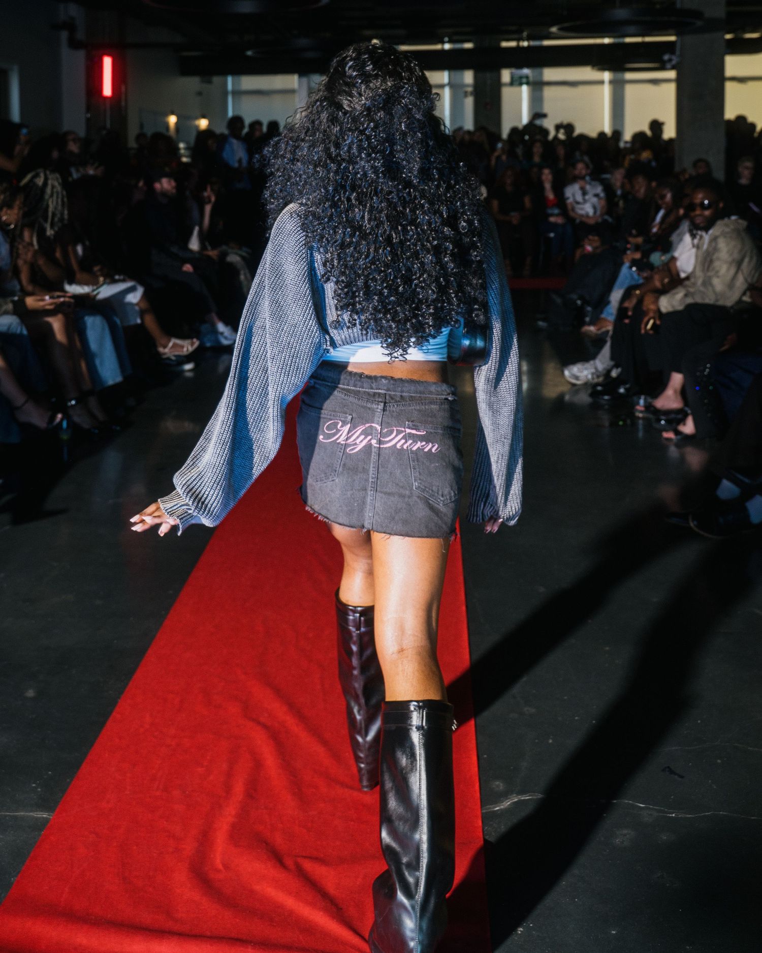 Rear view of model in denim jacket and My Turn branded mini skirt on red carpet runway.