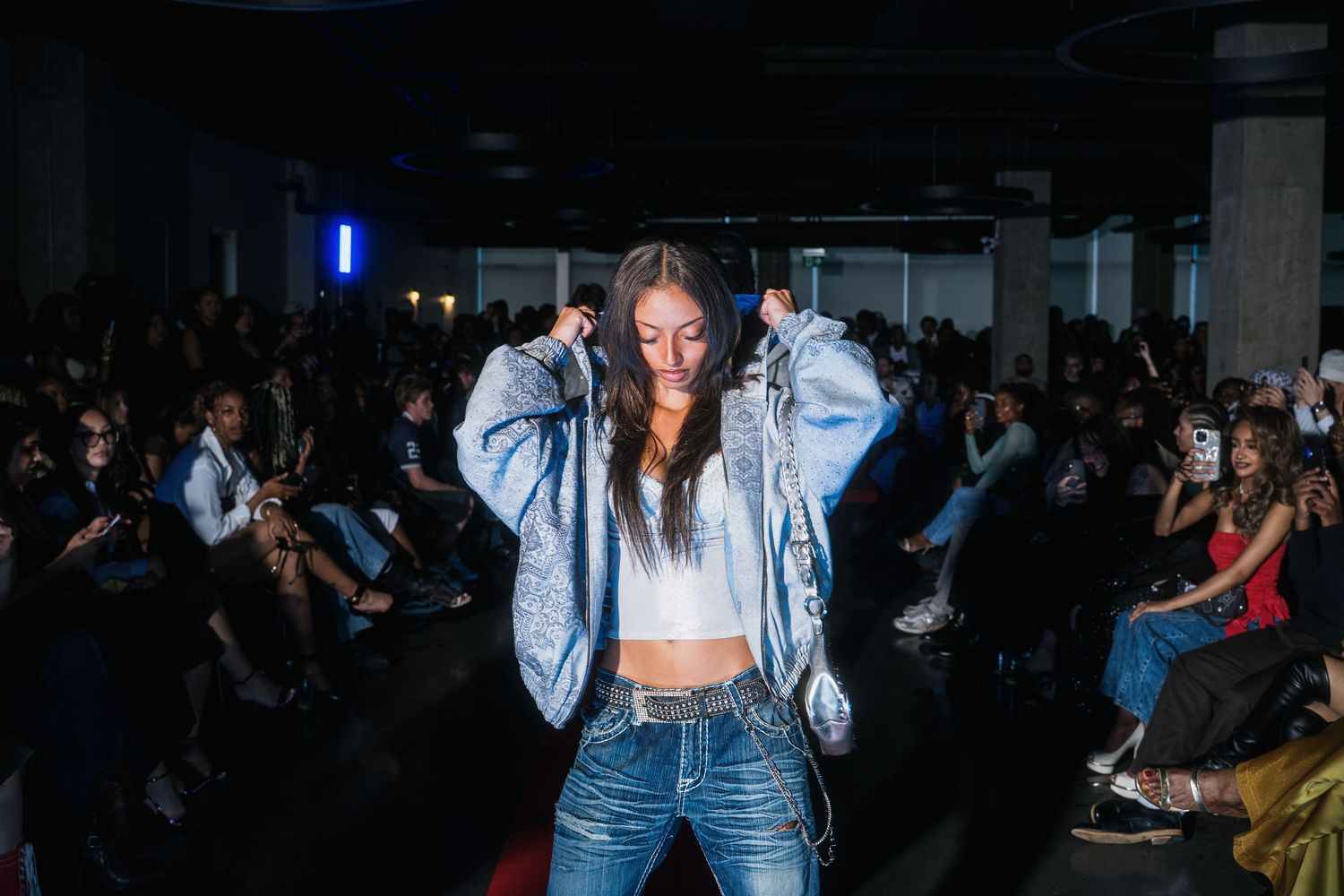 Female model adjusts silver denim jacket on runway wearing white crop top and jeans at fashion show.