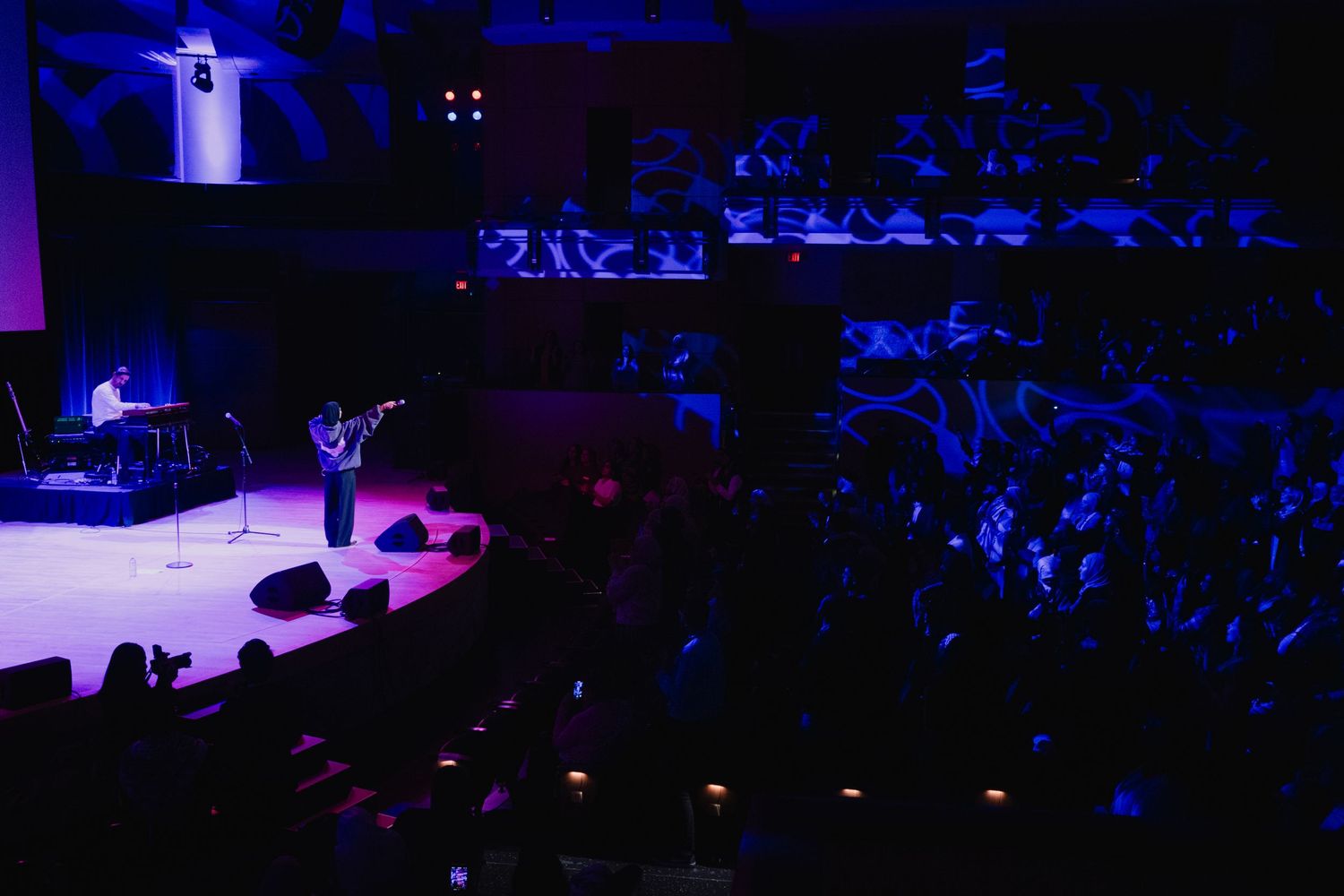 Performer on stage playing violin for a large crowd in a dramatic purple-lit concert hall.
