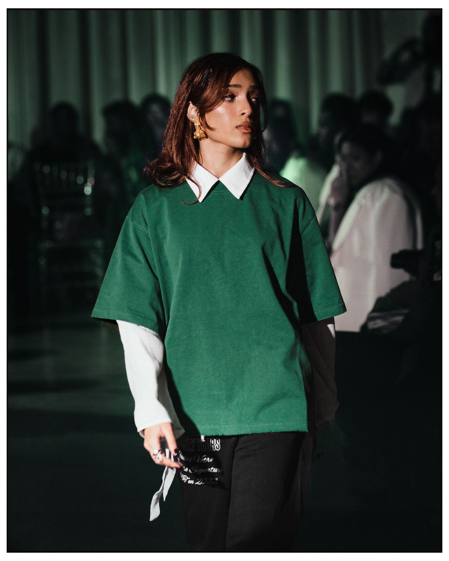 Model walks runway in oversized green t-shirt layered over white collared shirt with black pants.