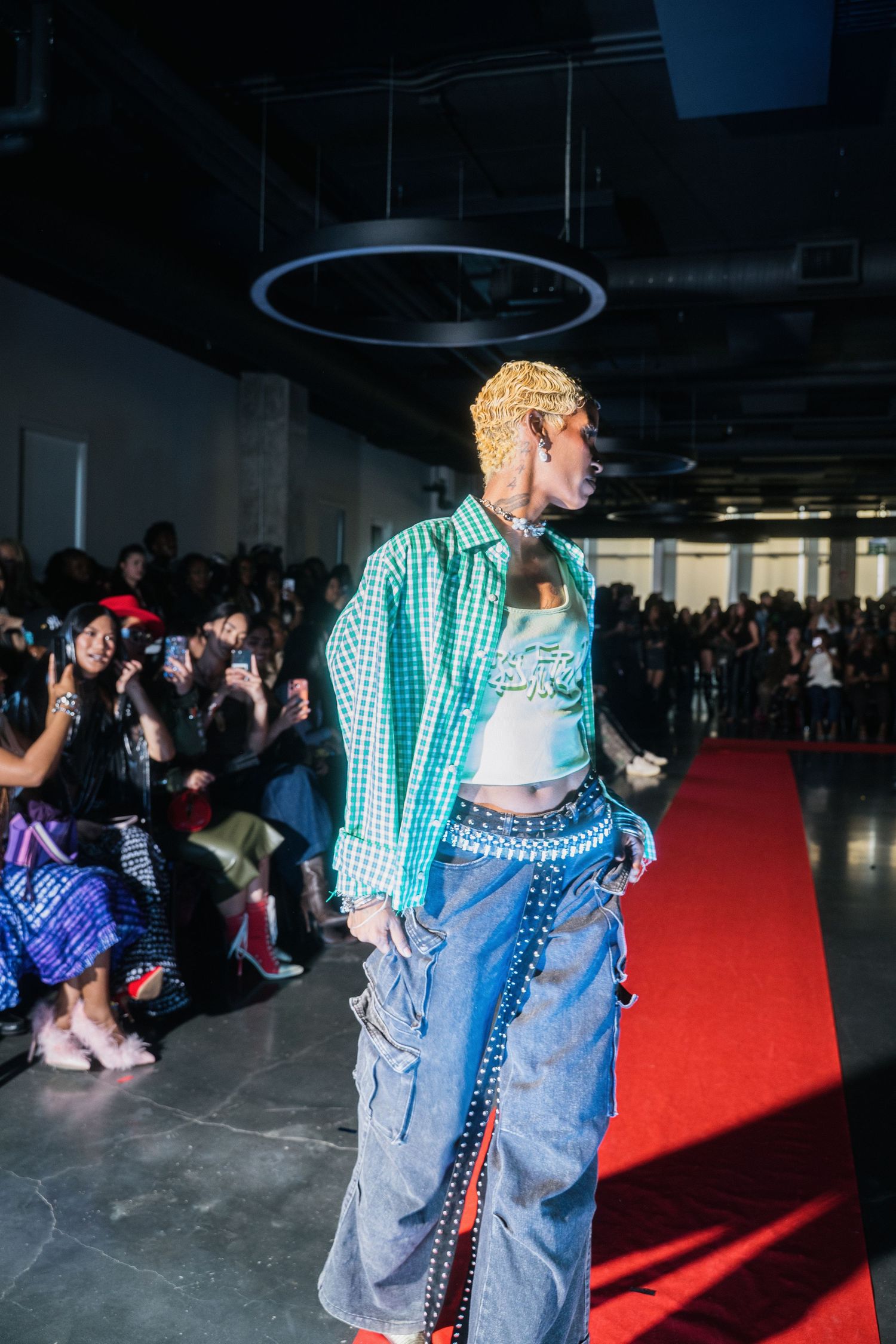 Model walks runway in green gingham jacket and studded denim at urban fashion show.