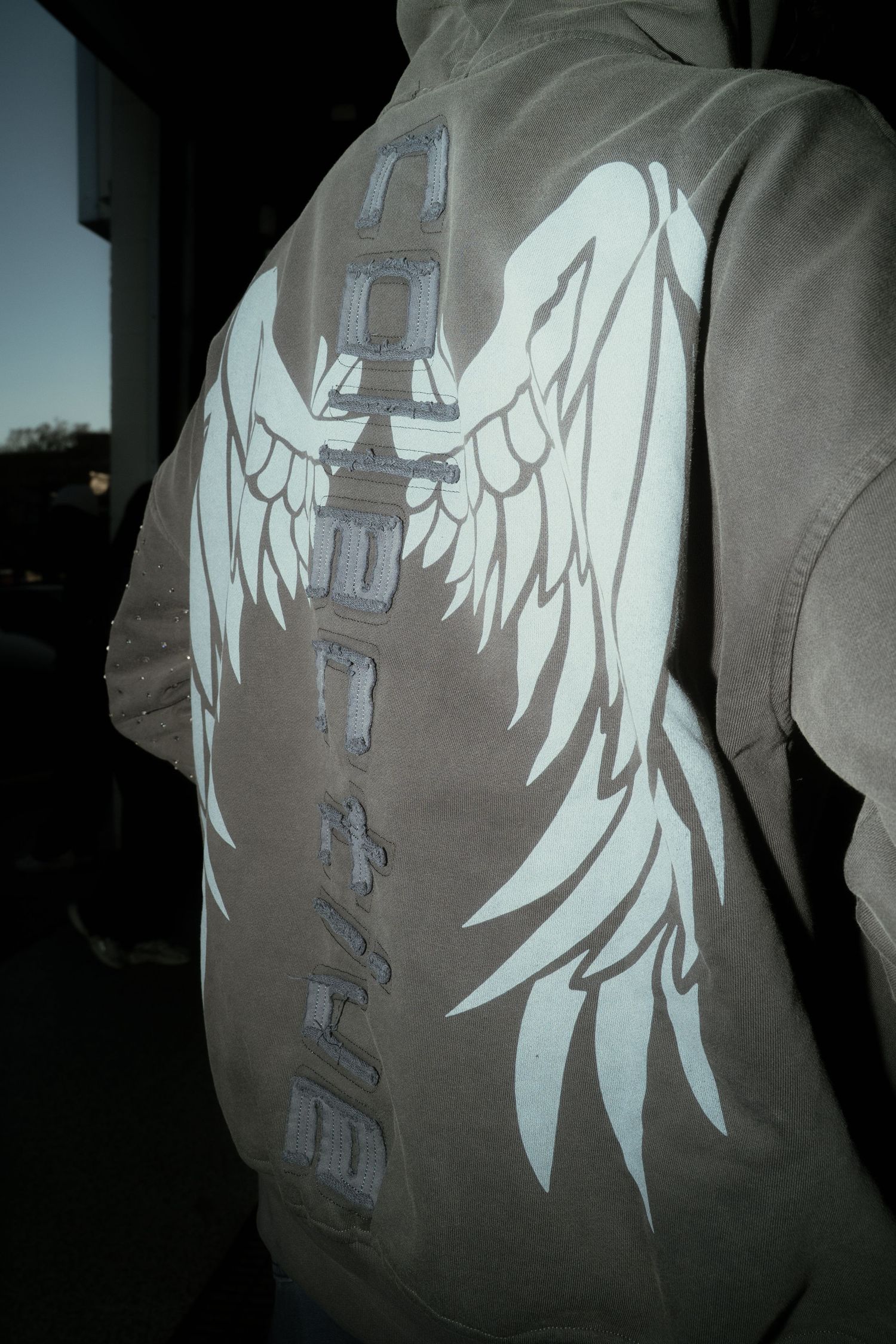 Back view of olive hoodie with white angel wings graphic and vertical text design.