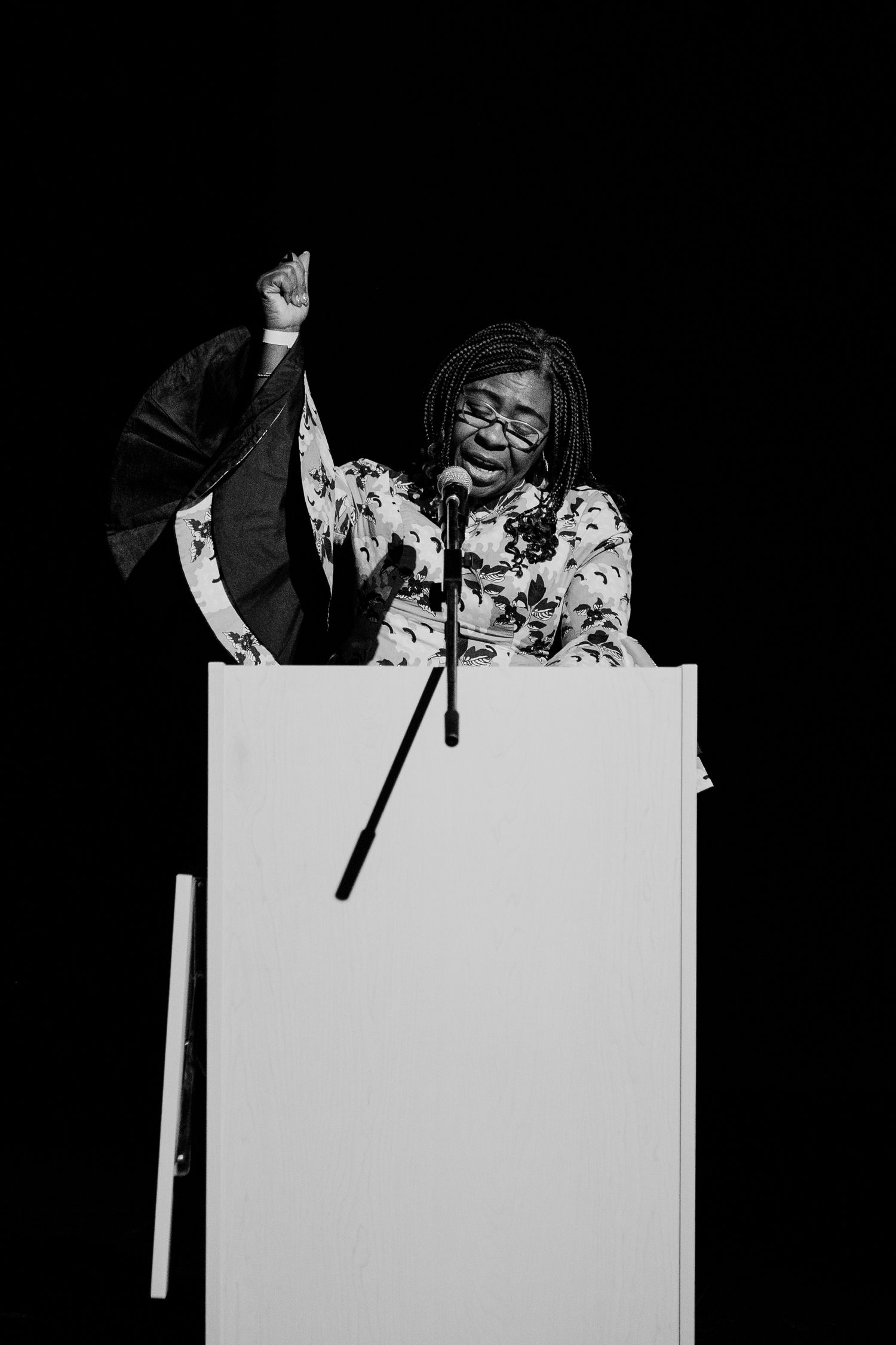 A woman raises her fist triumphantly while speaking passionately at a podium on a darkened stage.