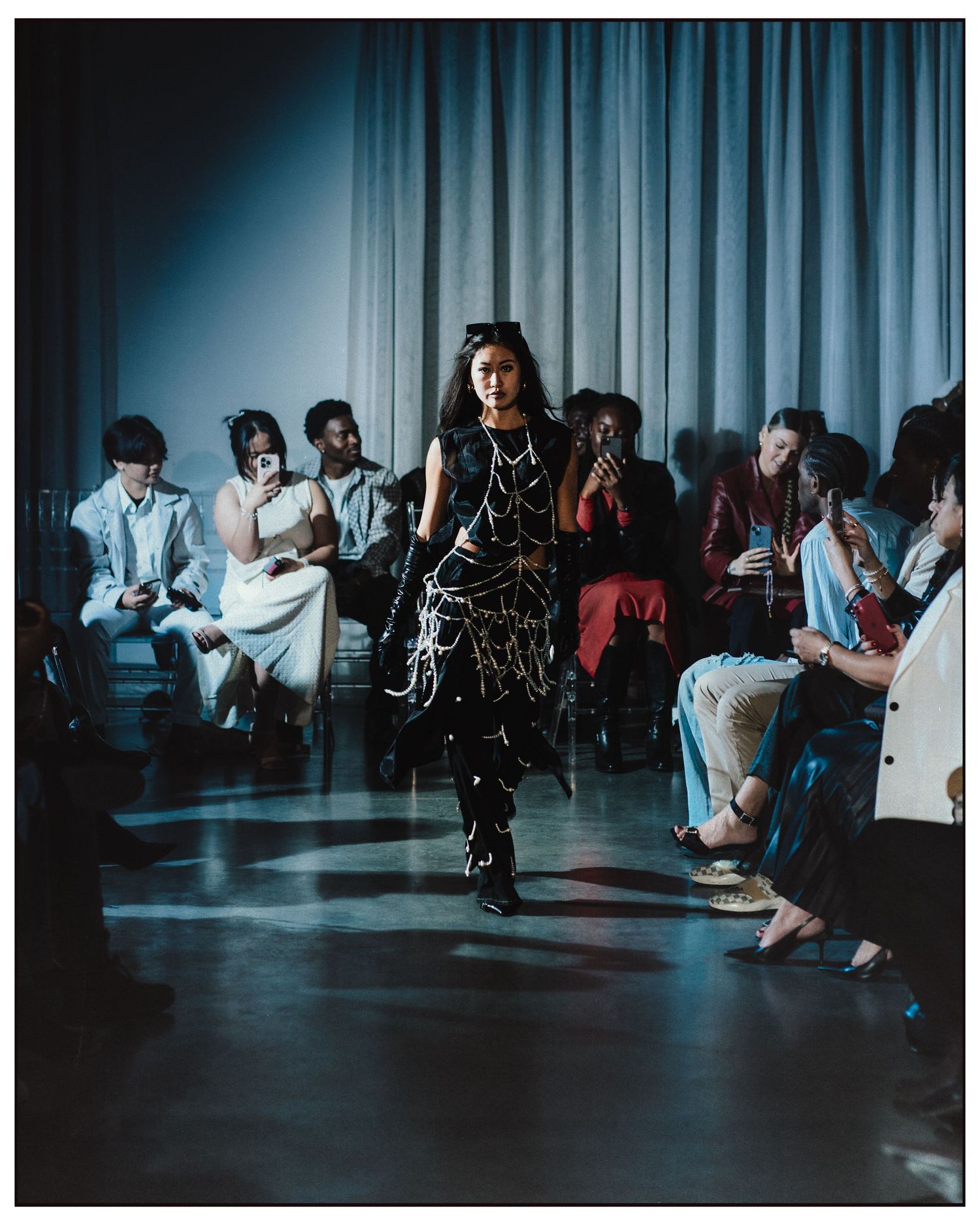 Model walks runway in black dress with white chain embellishments at indoor fashion show.