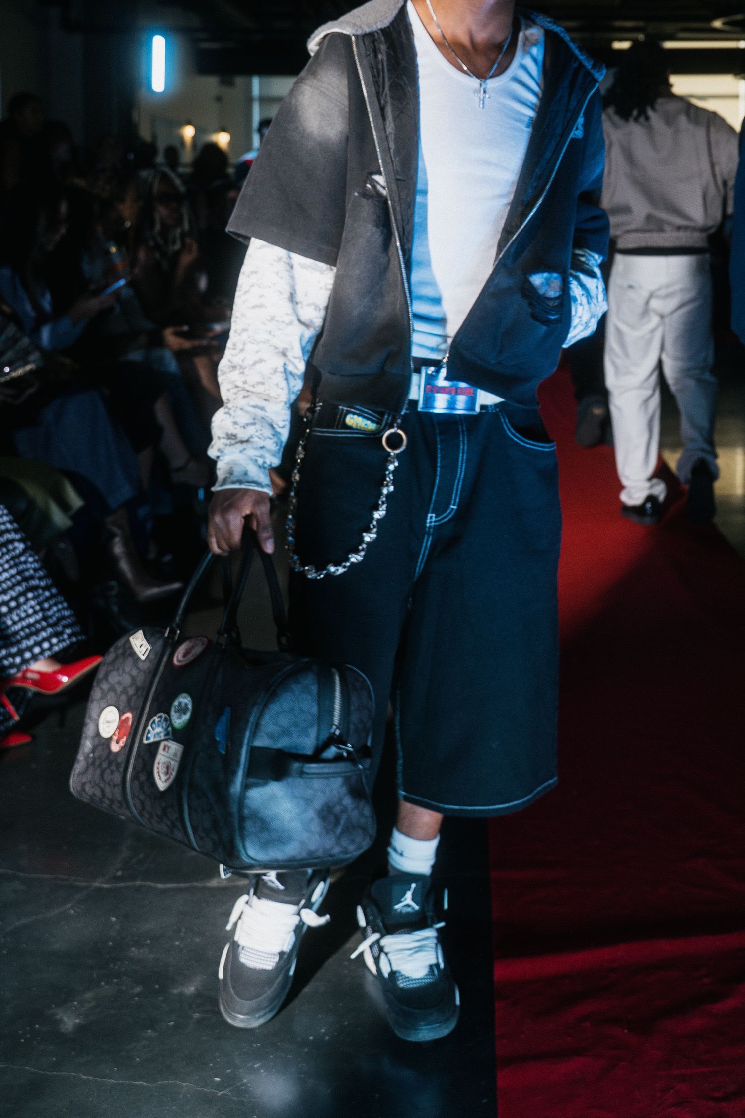 Detail shot of model in layered jacket with chain belt, carrying Coach bag, wearing Jordan sneakers.