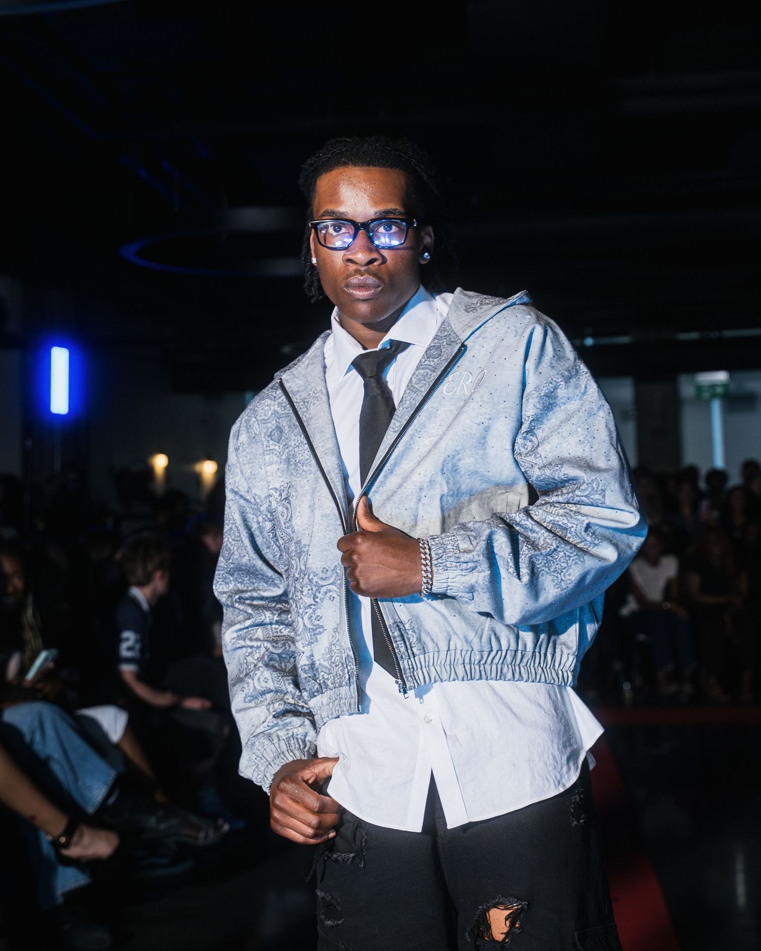 Male model walks runway in silver paisley bomber jacket with white shirt and black tie at fashion show.