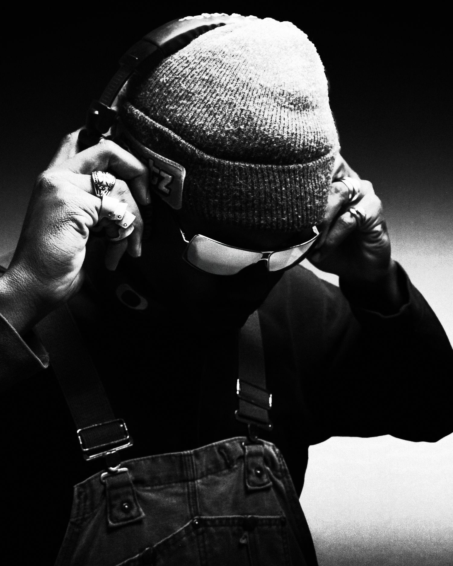Black and white photo of person adjusting headphones wearing beanie and overalls.