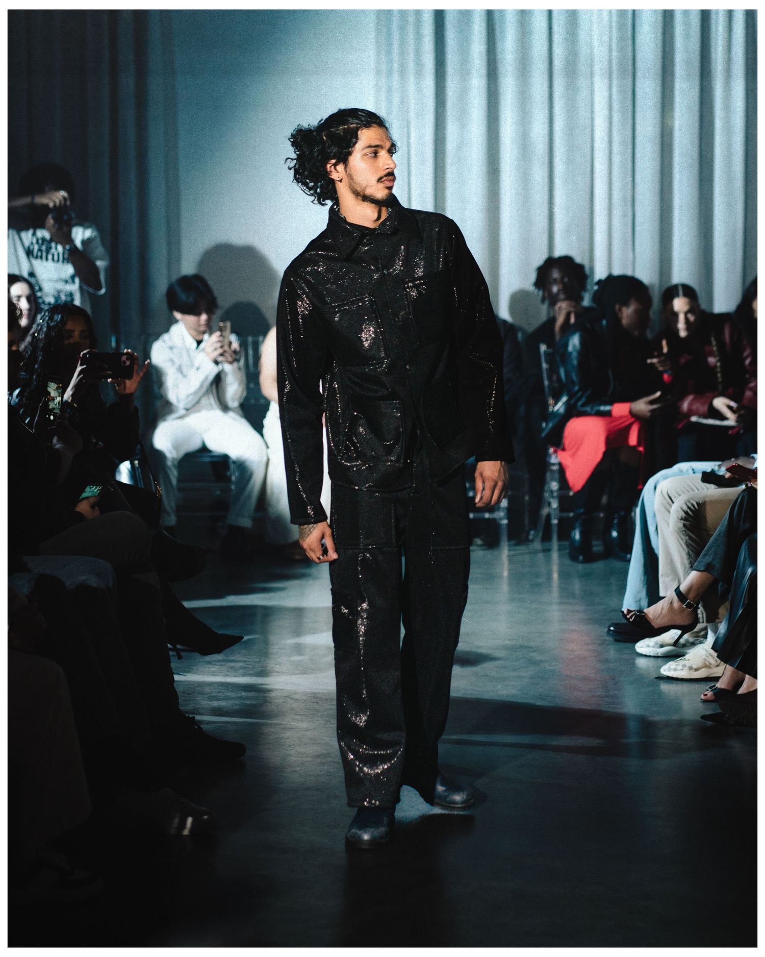 Male model walks runway in all-black sequined outfit at indoor fashion show.