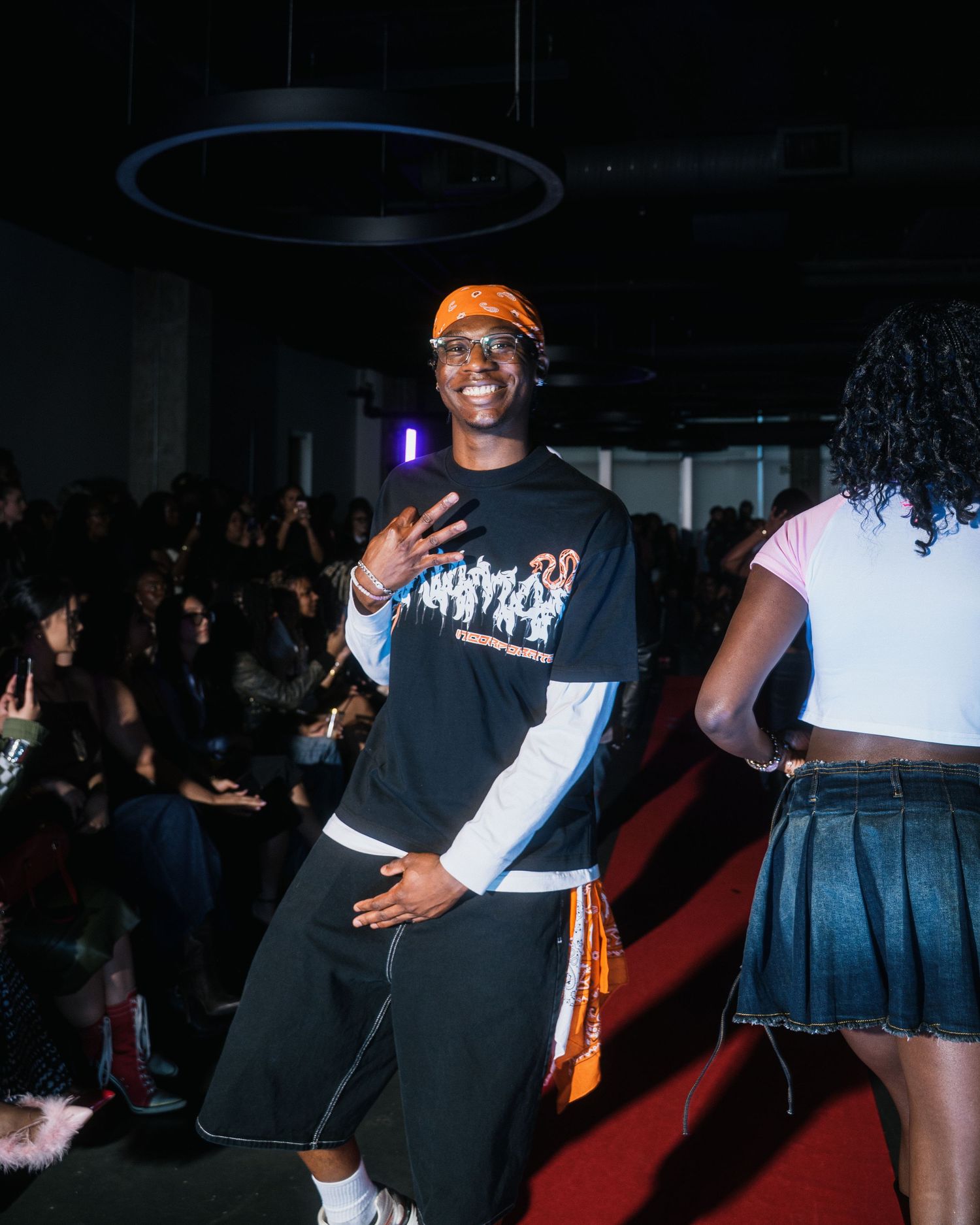 Smiling male model waves to crowd on red carpet runway wearing black graphic sweatshirt and orange cap.