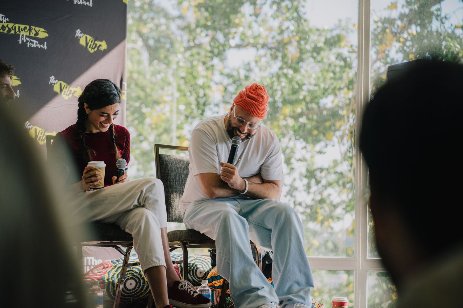 Two people sit and chat at an outdoor festival panel discussion, one holding a coffee cup and the other a microphone.