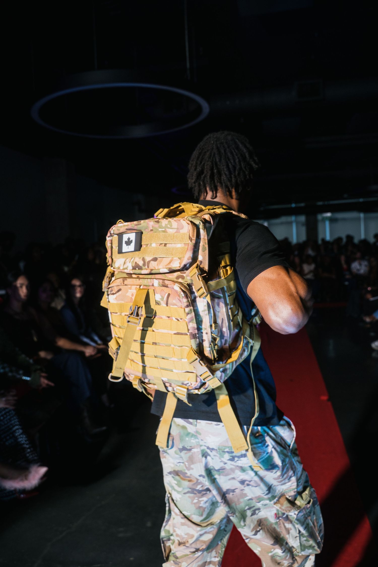Model walks runway wearing yellow camo tactical backpack and matching camo pants at urban fashion show.