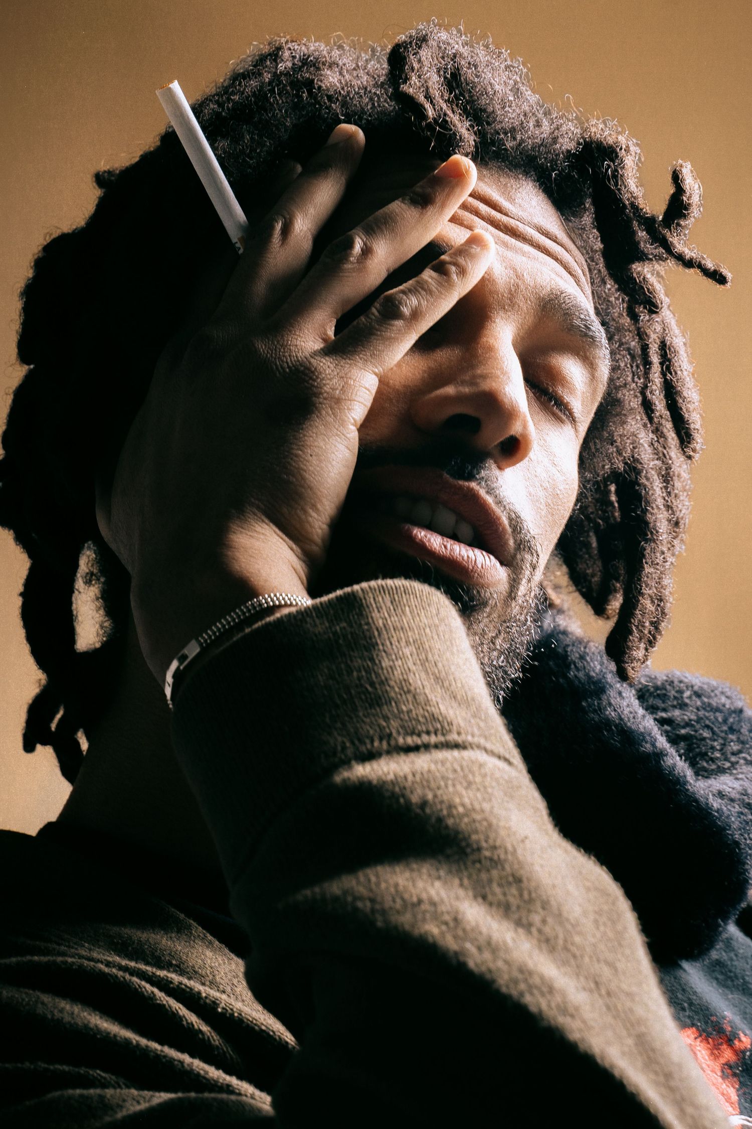 Man with dreadlocks holding cigarette near face in warm-toned dramatic portrait.