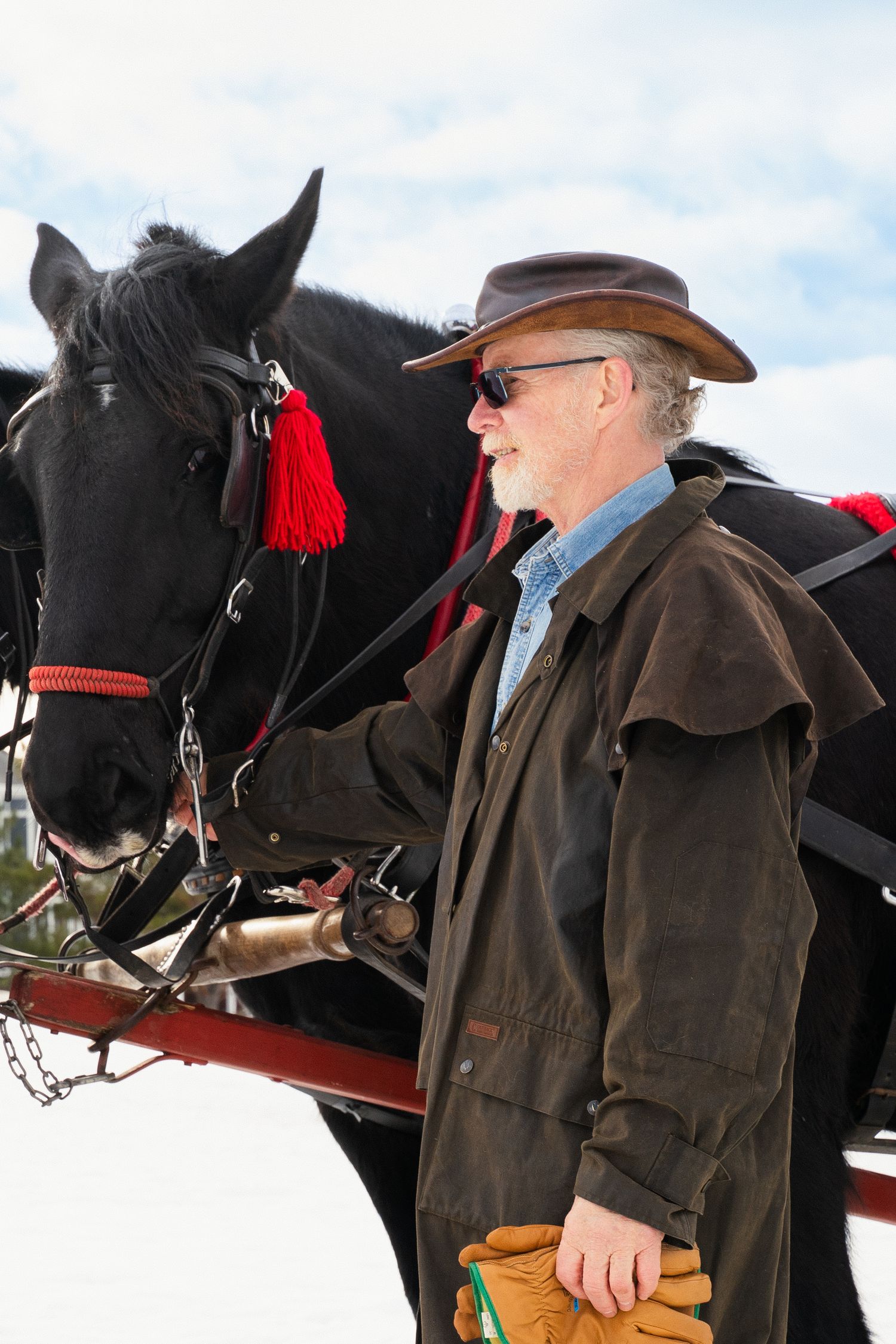 A cowboy in a brown hat and coat stands beside a harnessed black horse with red tassels outdoors.