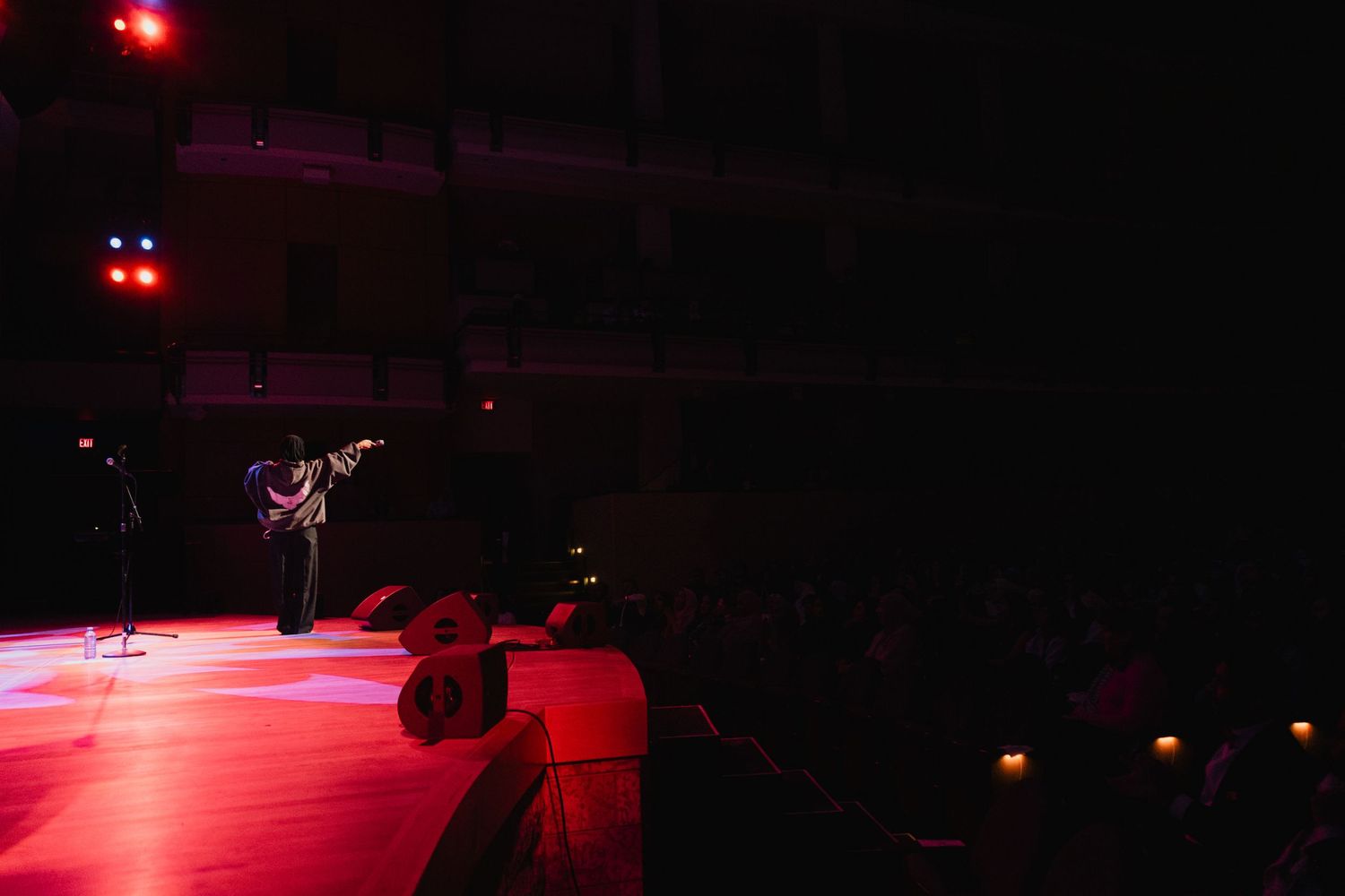 A lone guitarist performs on a red-lit stage in a large darkened concert venue.