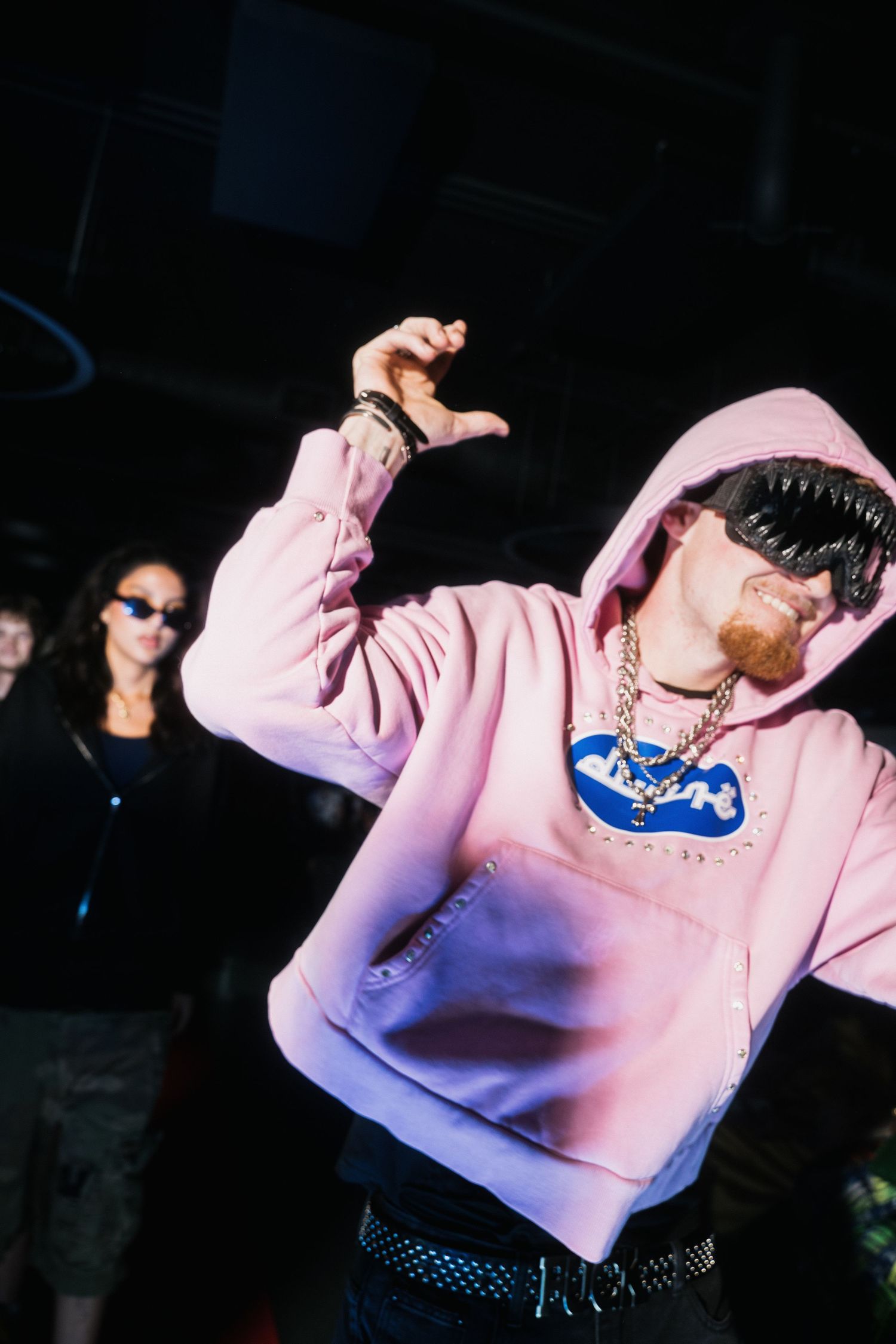 Man in pink hoodie with gold chain dances energetically at a dark nightclub event.