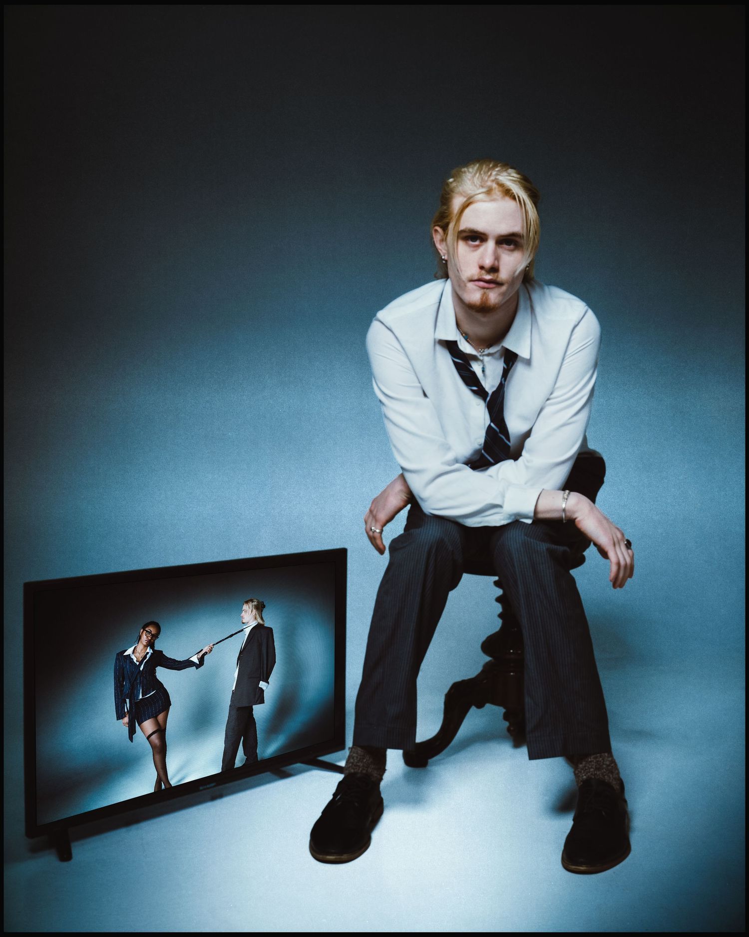 Blonde young man in white shirt and tie sits beside a TV screen displaying a couple in a studio.