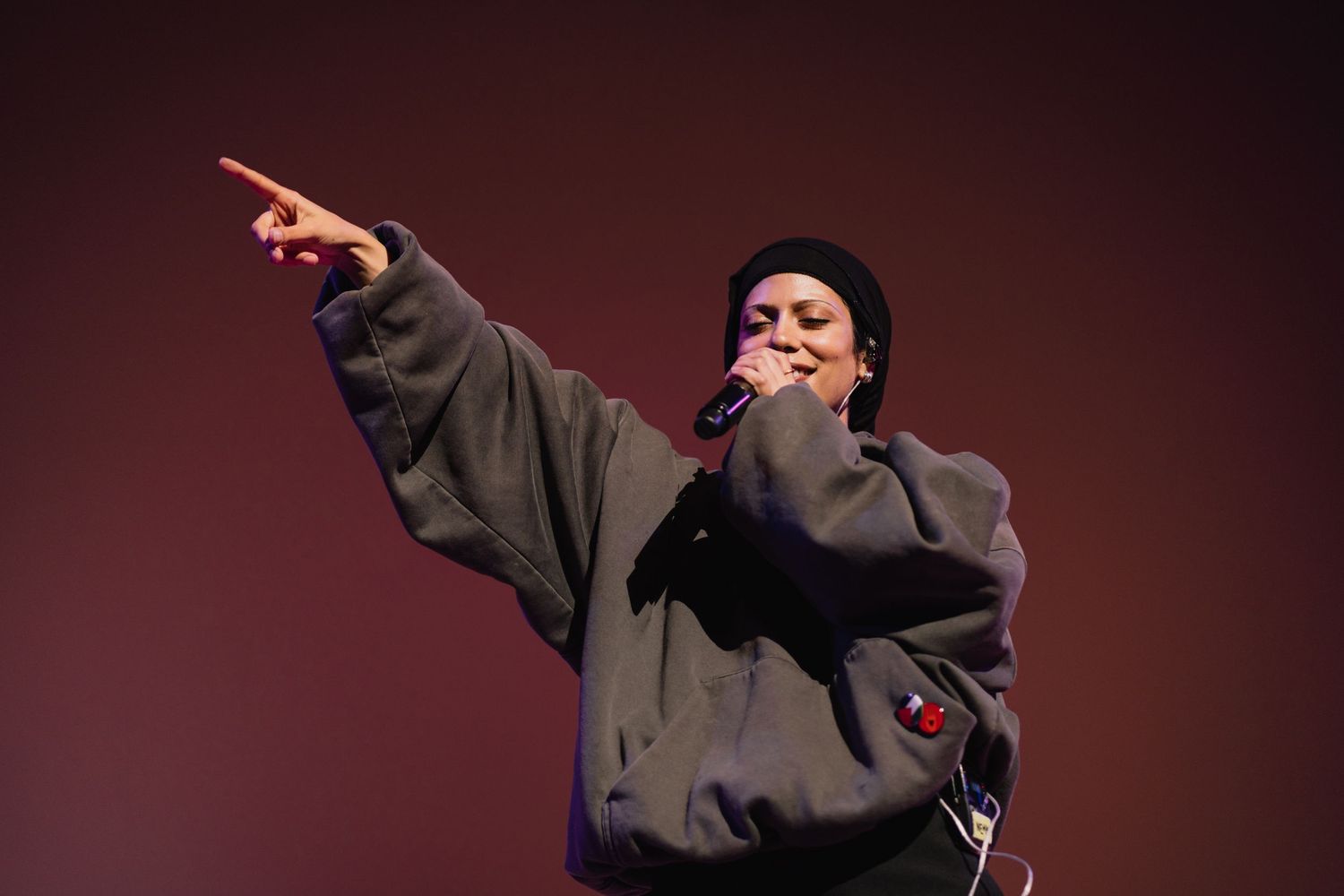 A performer in a gray hoodie and black beanie sings into a microphone while pointing at the crowd.