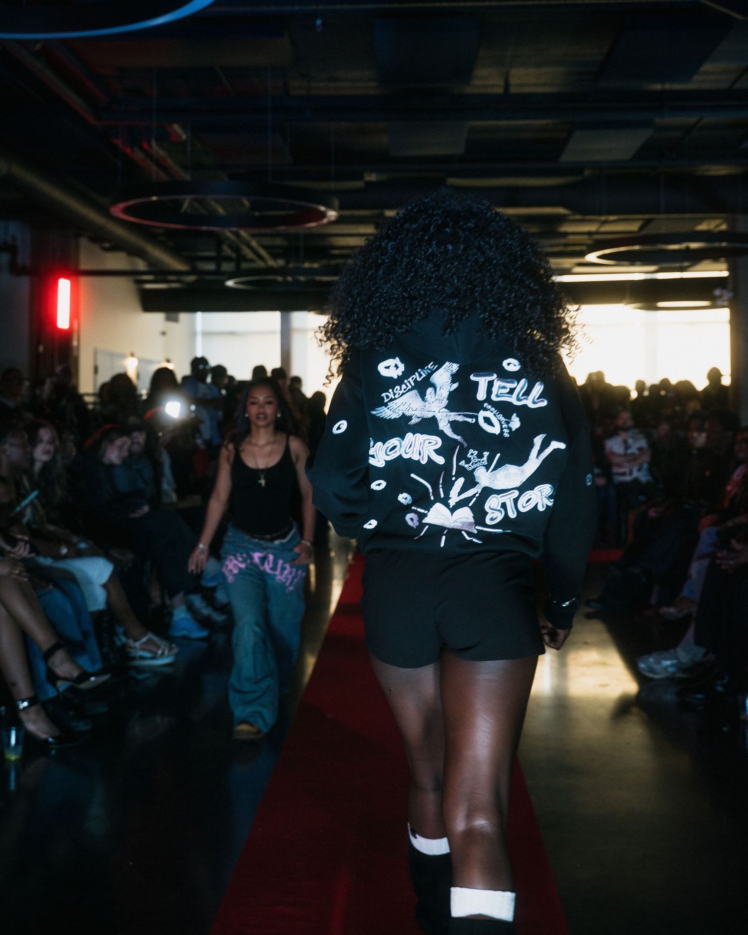 Model walks runway in black graphic jacket reading 'Tell Your Story' at urban streetwear fashion show.
