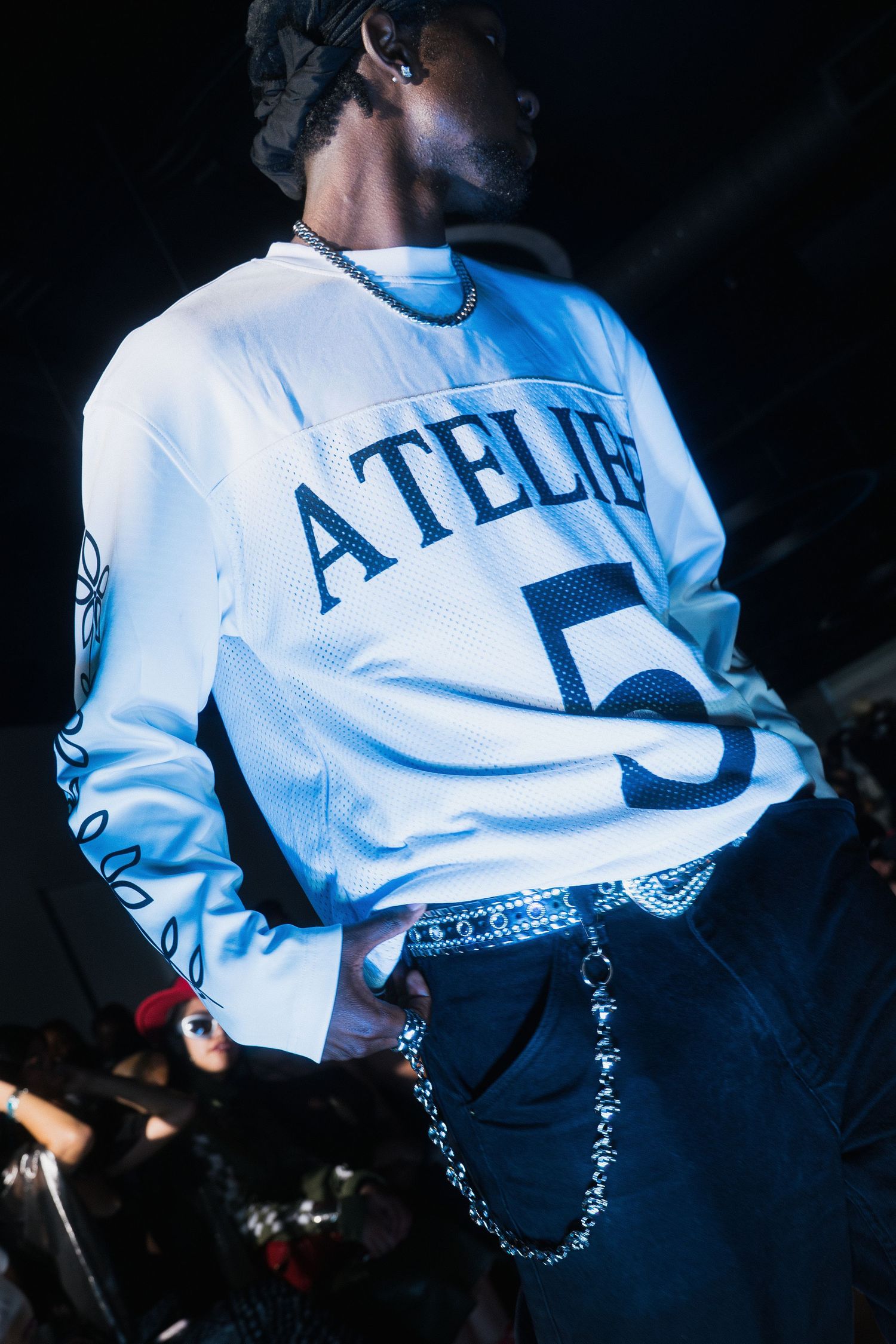 Male model wearing a white Atelier Five jersey with chain accessories under blue stage lighting.