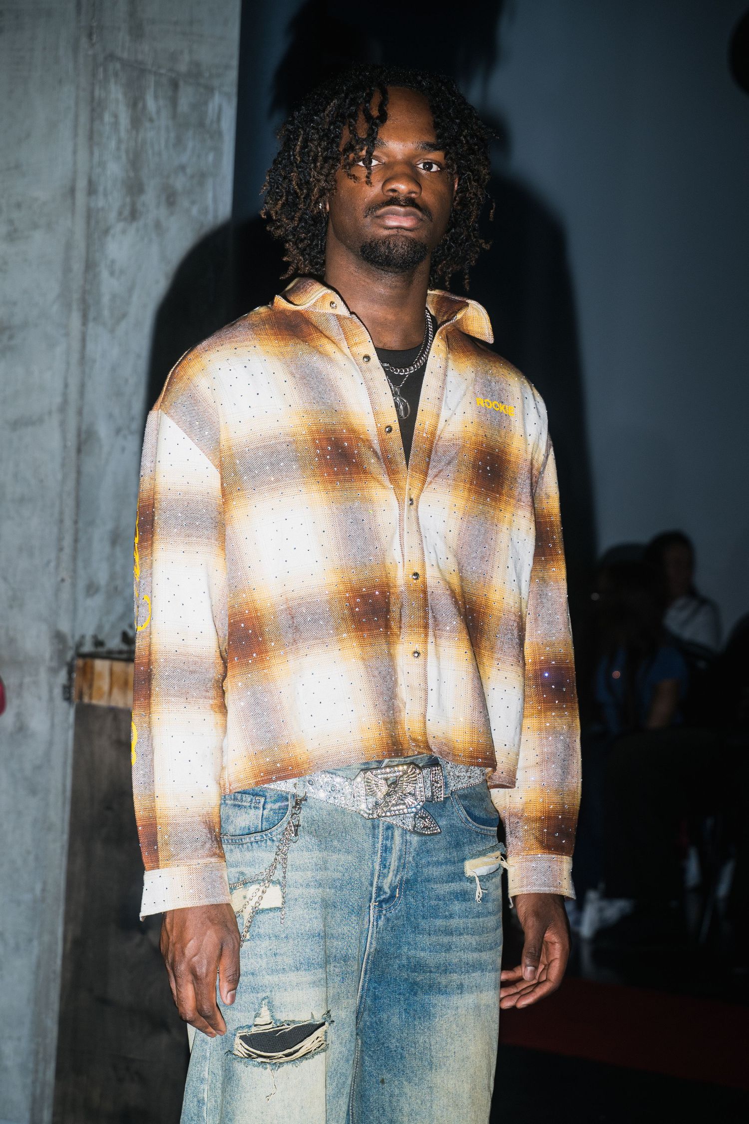 Male model with dreadlocks wearing plaid flannel shirt and distressed denim jeans at fashion show.
