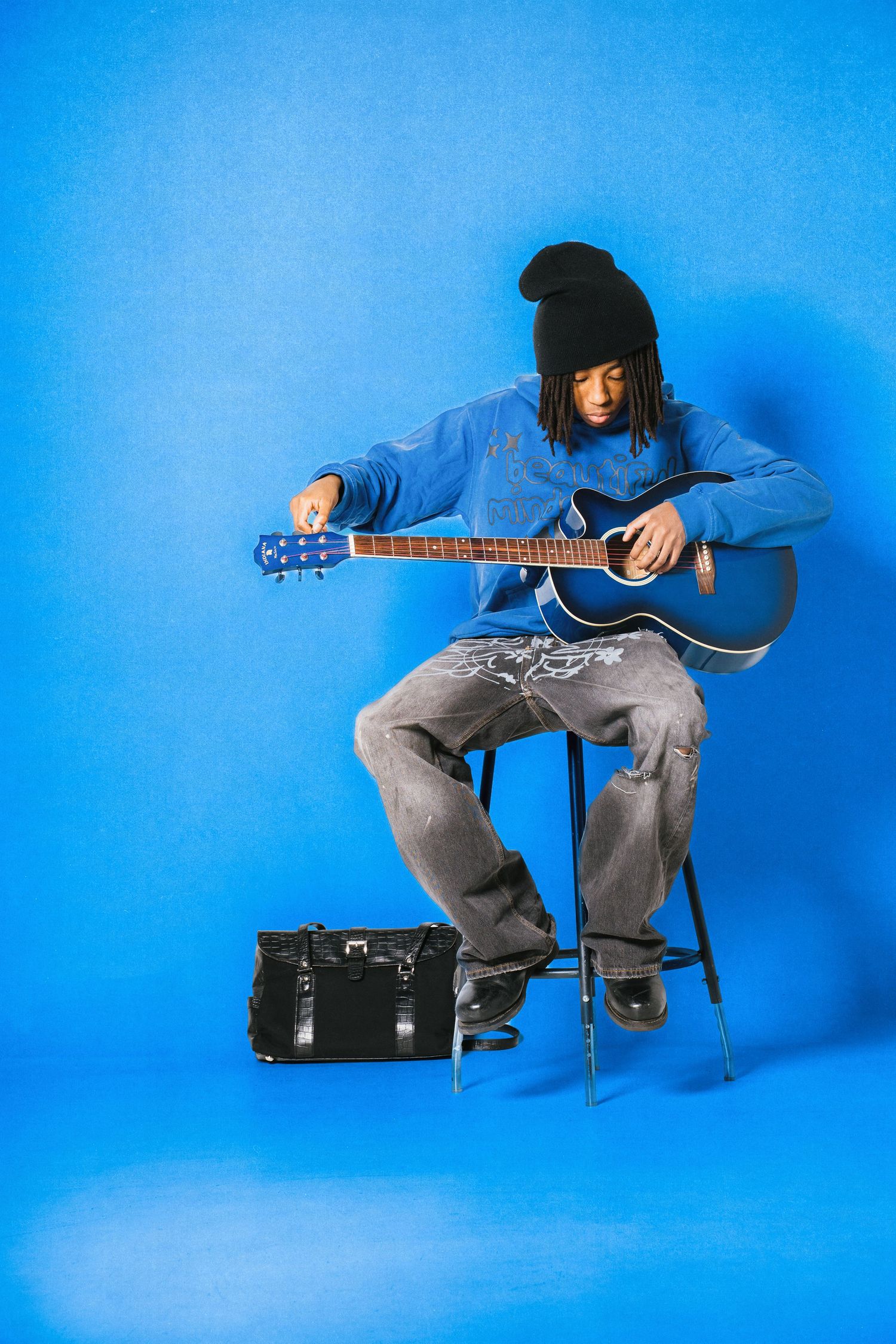 Young musician in blue hoodie and black beanie playing acoustic guitar seated on stool against blue background.