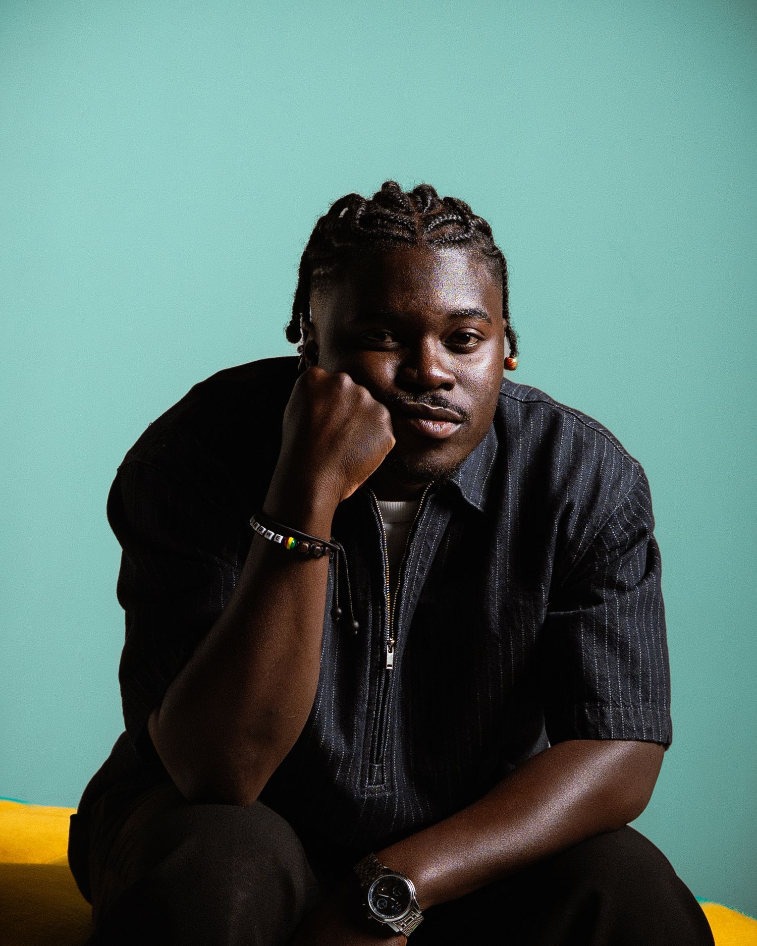 Young man in a black striped shirt sitting against a teal background, resting chin on hand.