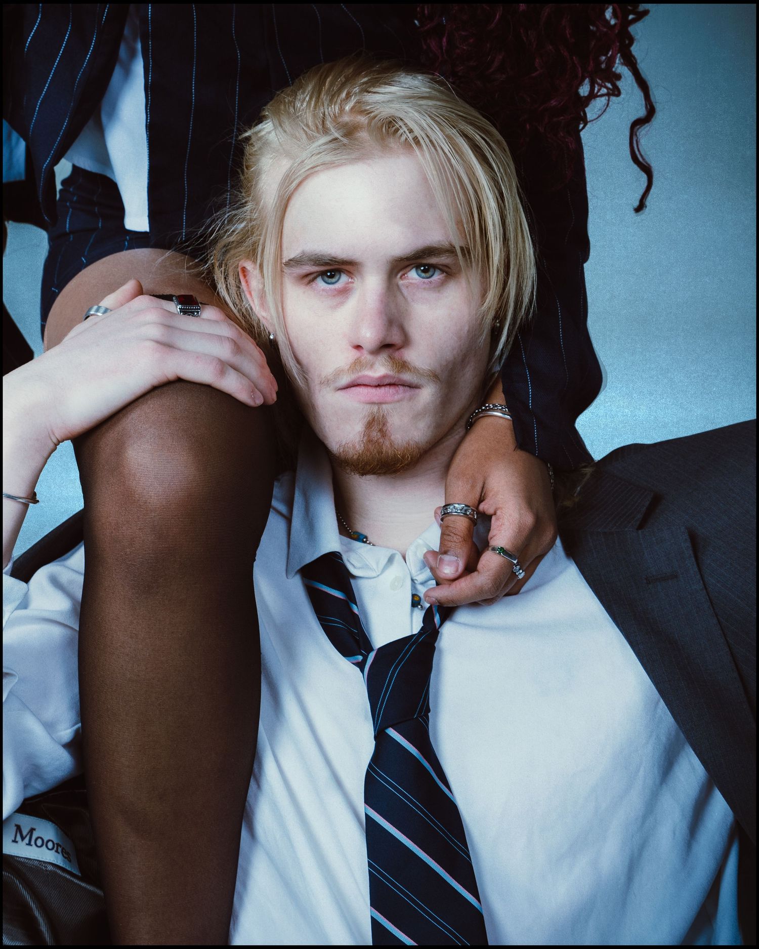 Blonde young man in a white shirt and tie reclines between two people in a blue studio setting.