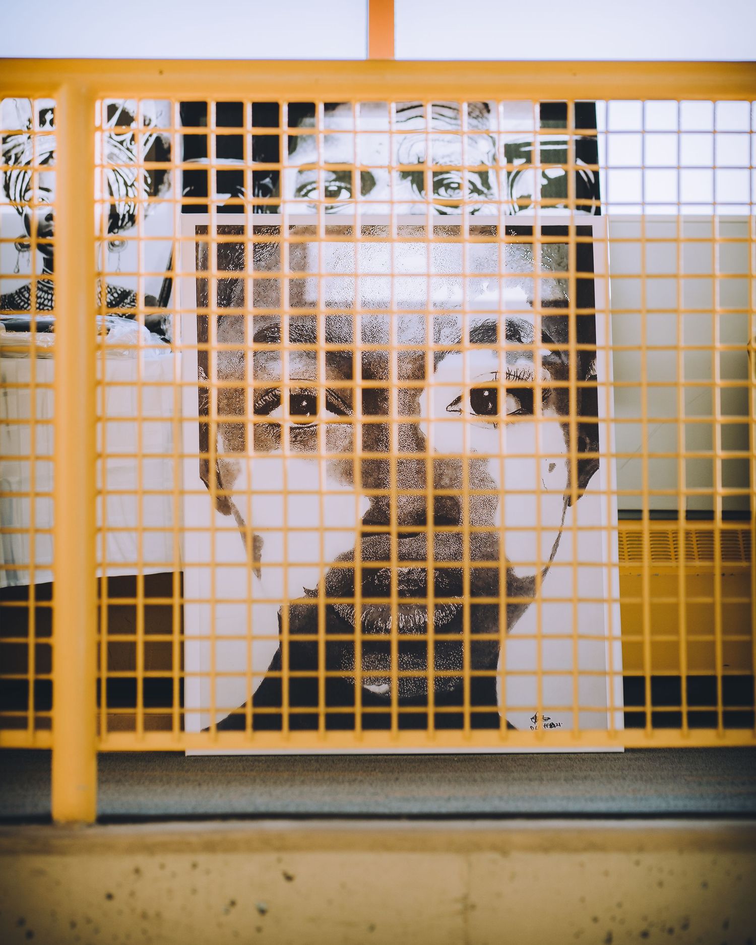 Black and white portrait artwork displayed behind yellow metal grid fence outdoors.