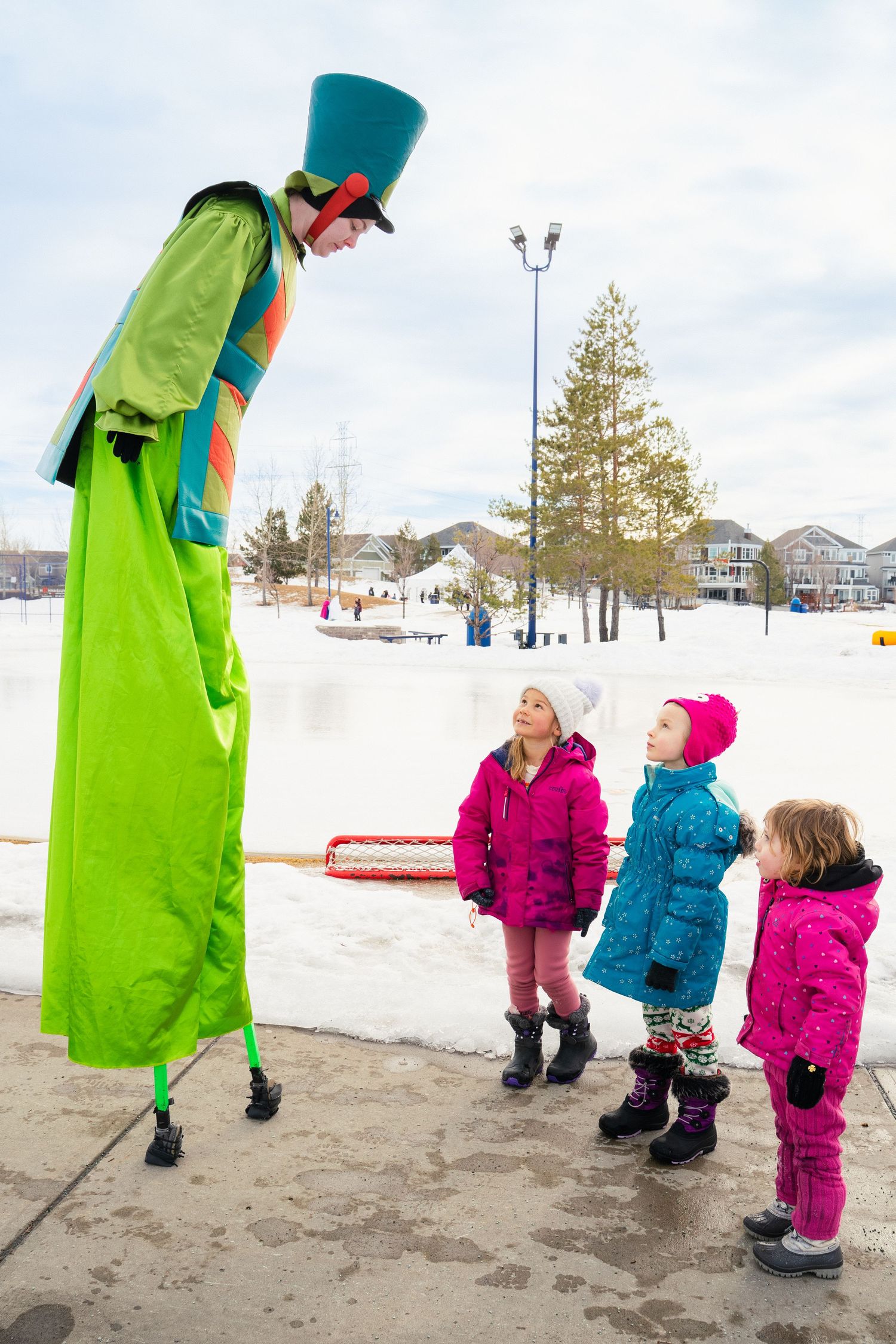 Stilt walker in colorful green costume entertaining three young girls in pink jackets at winter festival.