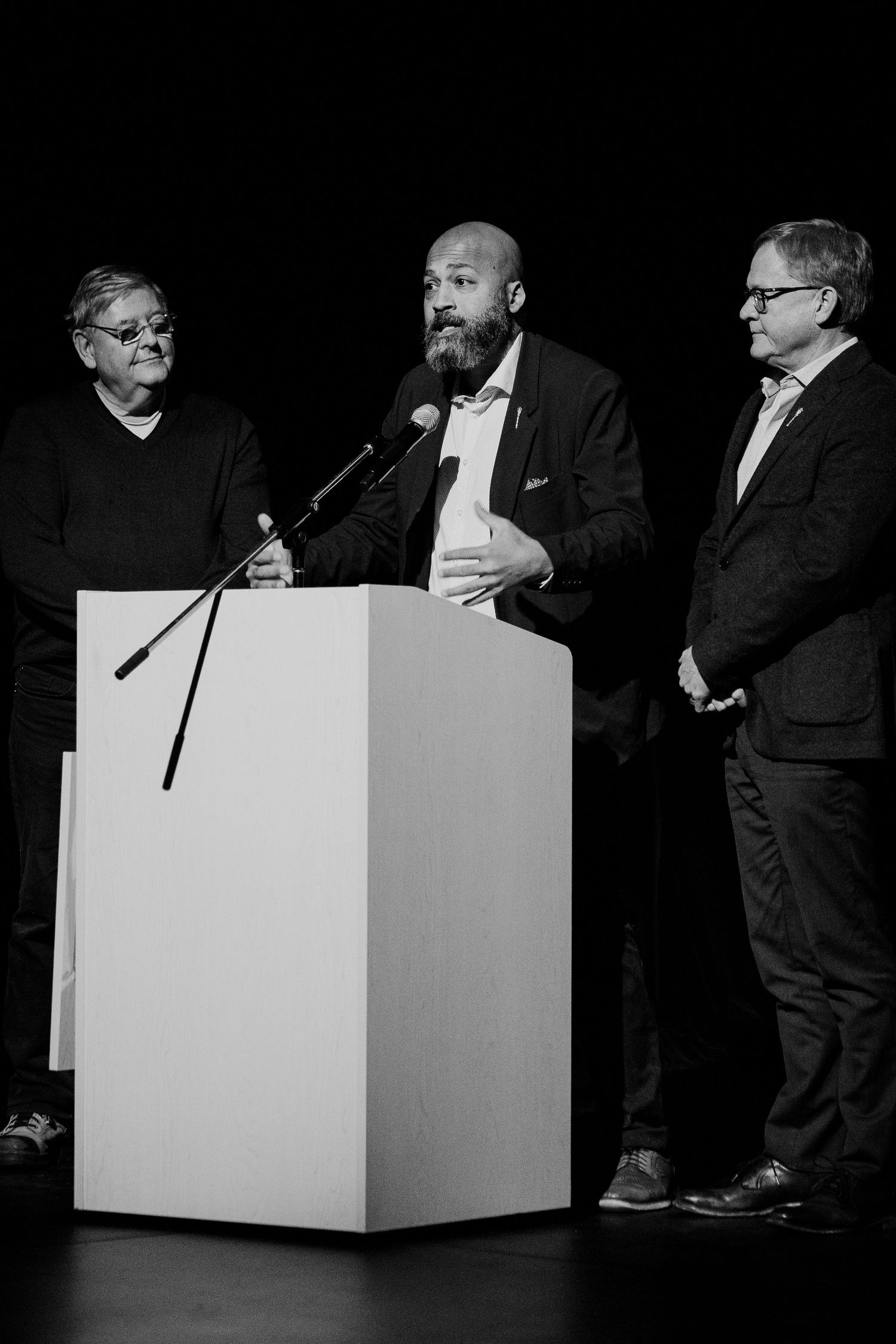 Three men stand at a podium on a dark stage, with the center man speaking into a microphone.