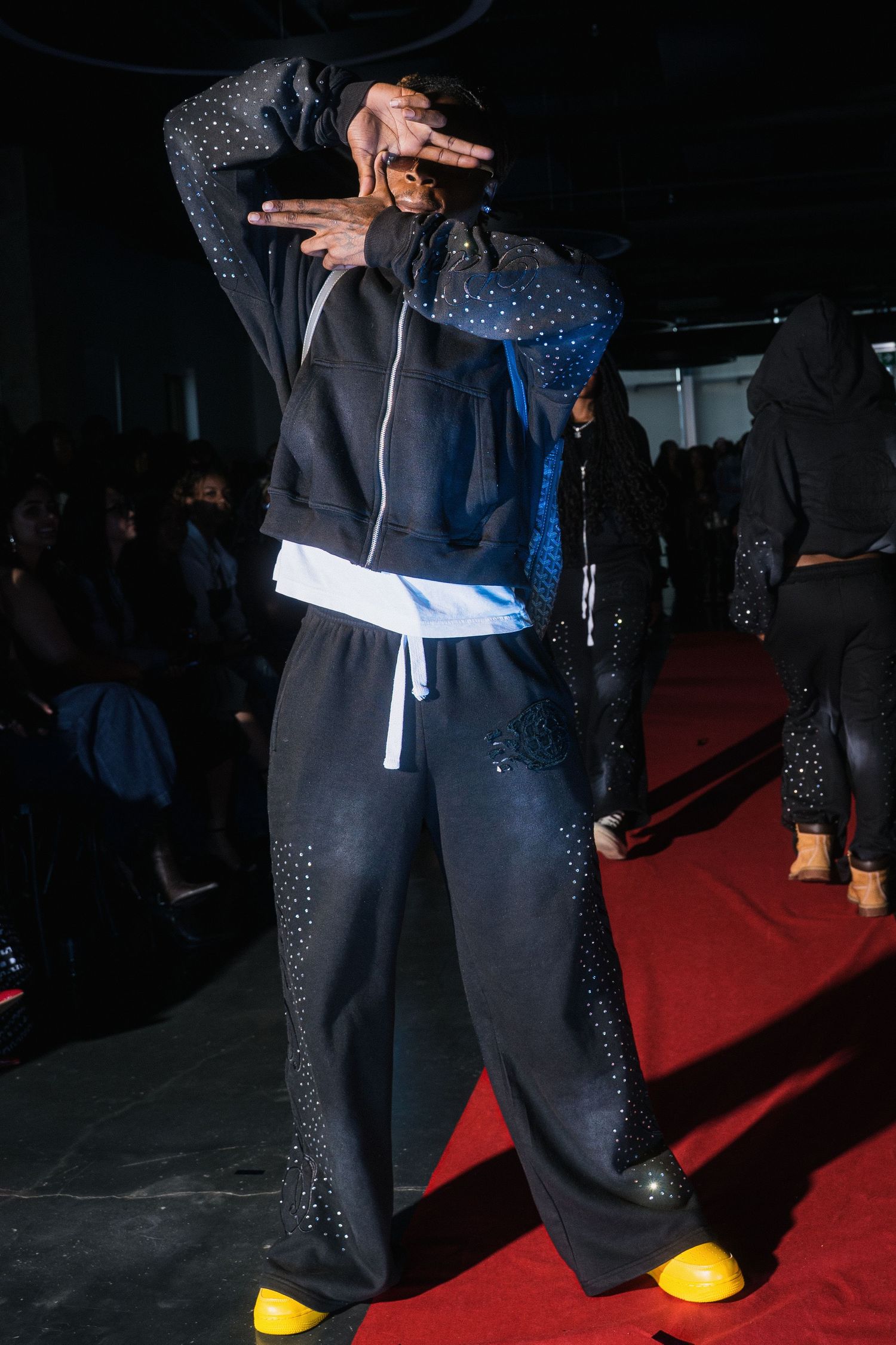 Model poses on red carpet runway in black rhinestone tracksuit with yellow shoes at fashion show.