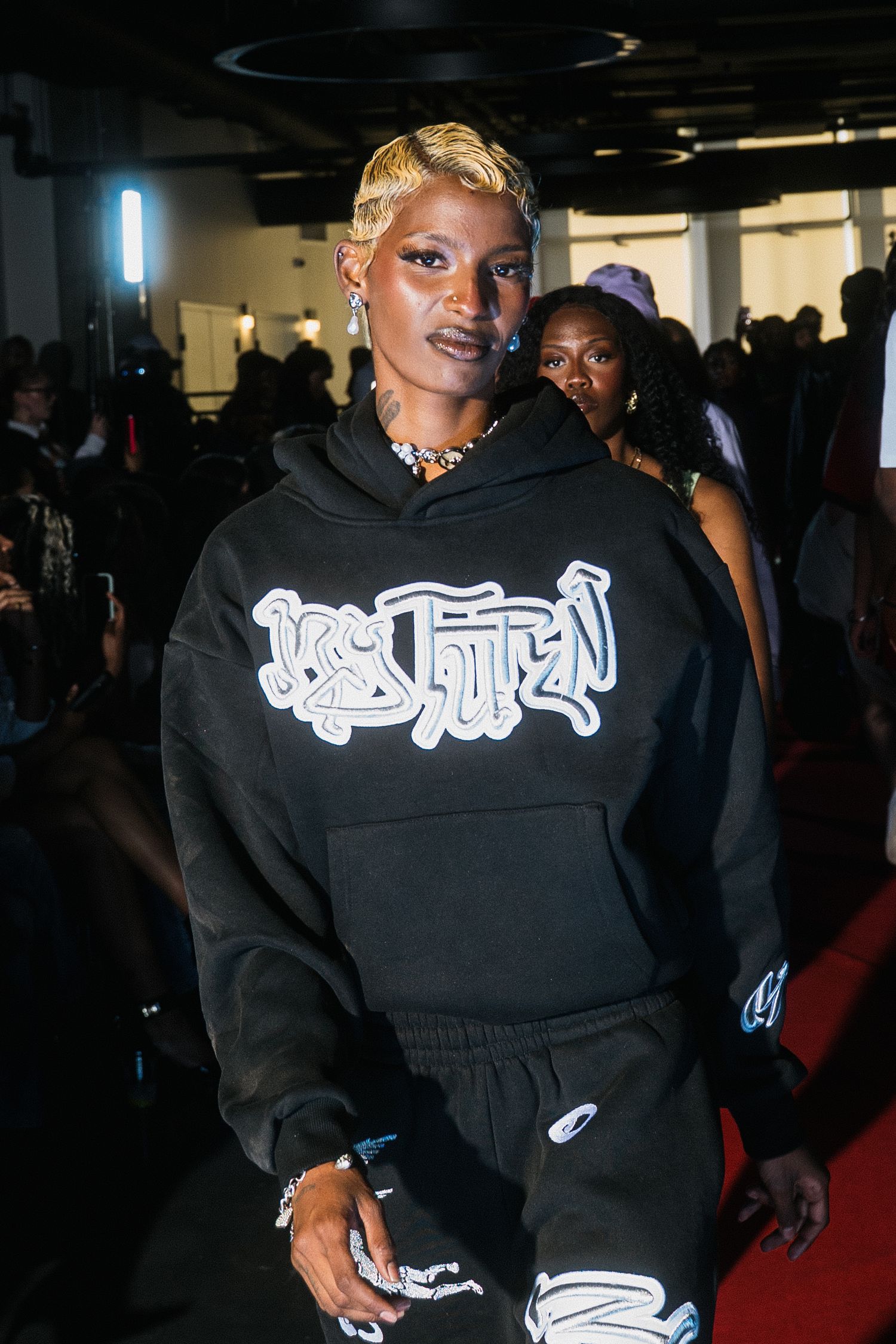Model wears oversized black hoodie with bold graffiti logo print at urban streetwear fashion event.