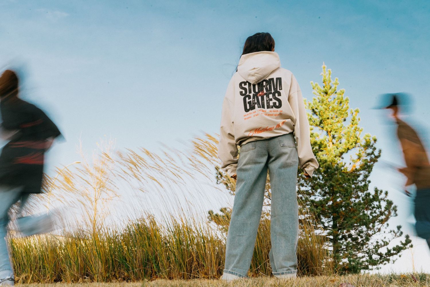 Person wearing Storm Gates hoodie standing outdoors with blurred figures and trees.